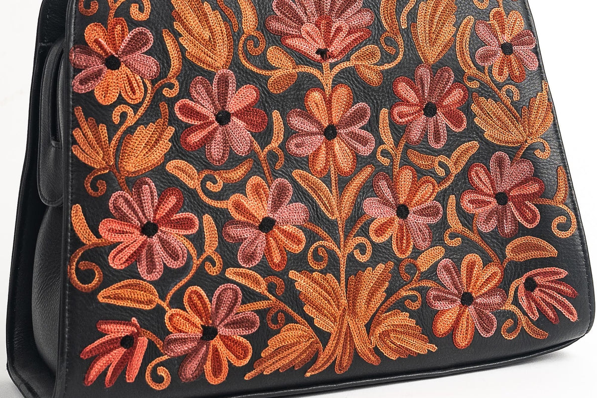 Close-up of a black handbag front with symmetrical coral-pink flowers, orange leaves and curling stems