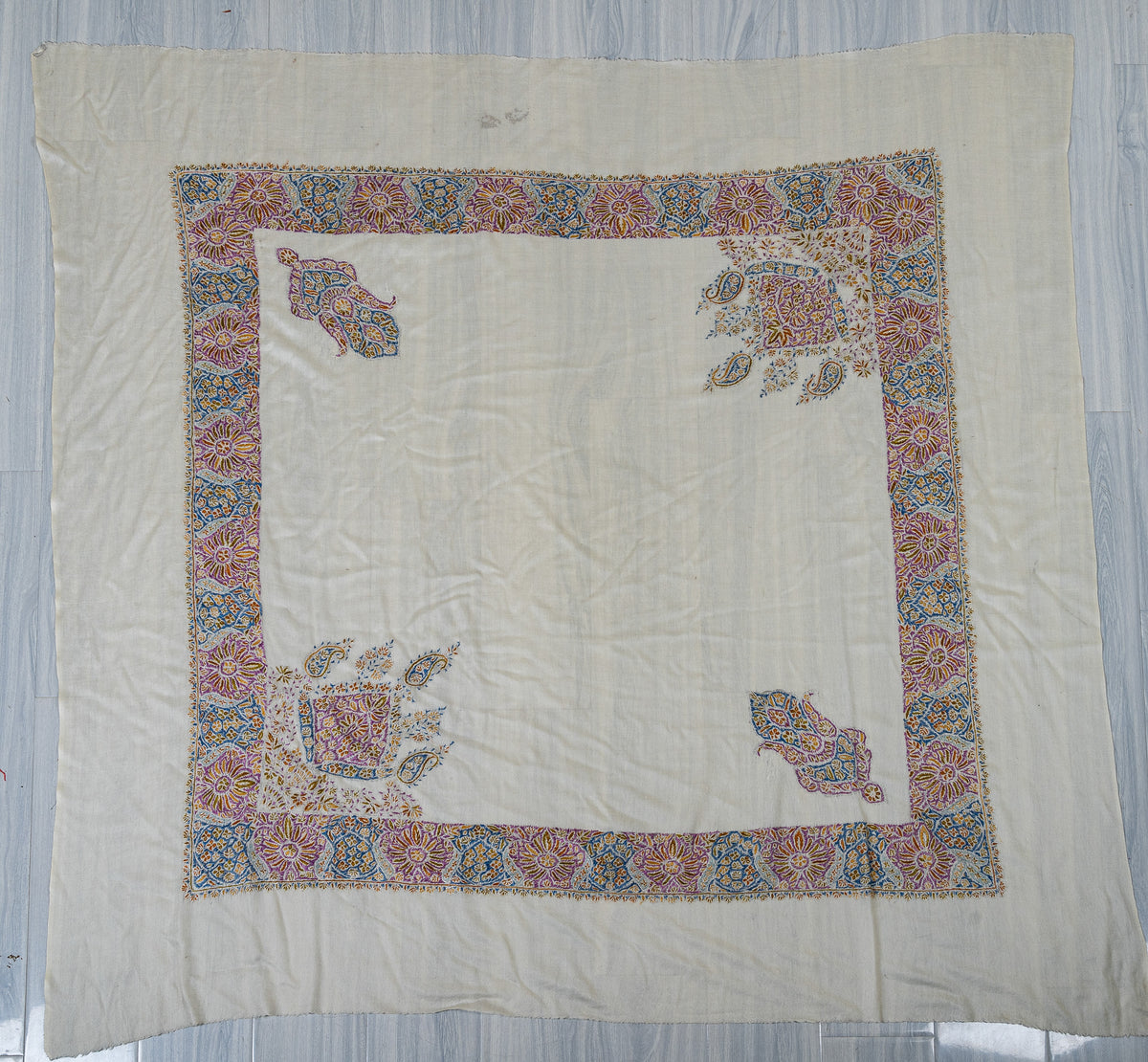 Ivory square Merino wool rumal laid flat, with a purple, blue and gold floral border and four embroidered corner motifs