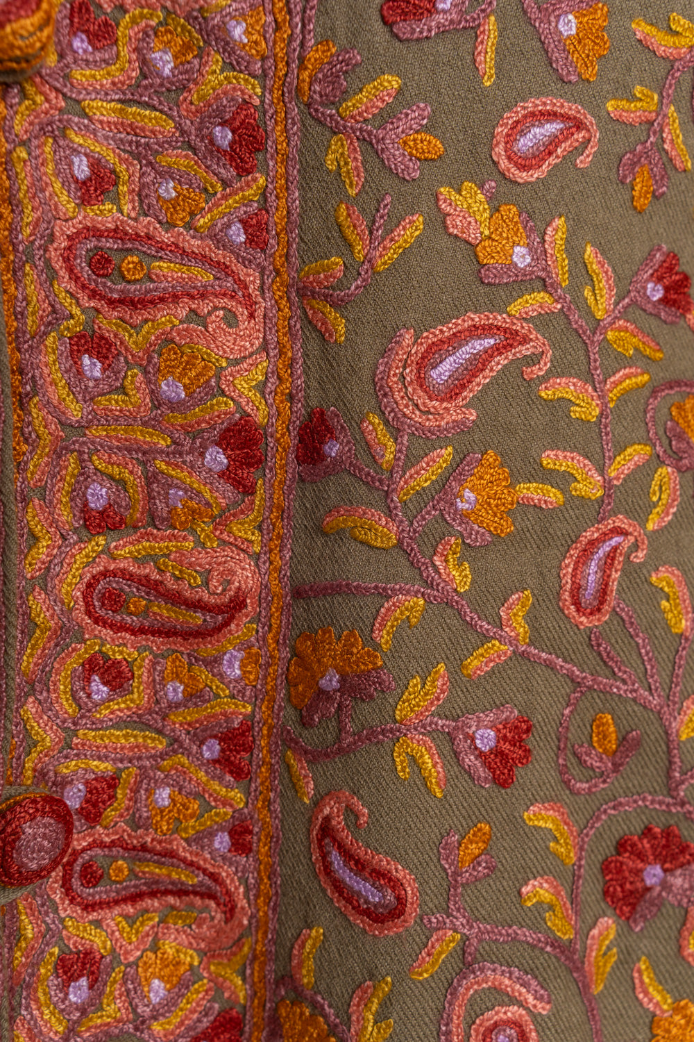 Close-up of raised pink paisleys, red flowers and orange Aari leaves beside a broad embroidered border on a brown jacket