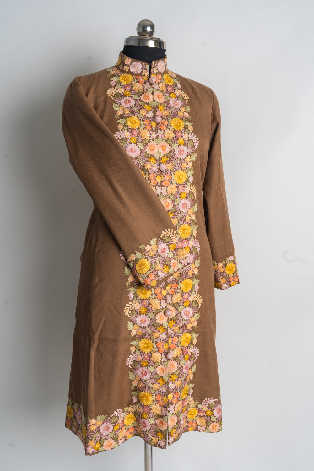 Full front of the brown long jacket on a mannequin, with one sleeve folded inward and floral embroidery along the opening and hem.