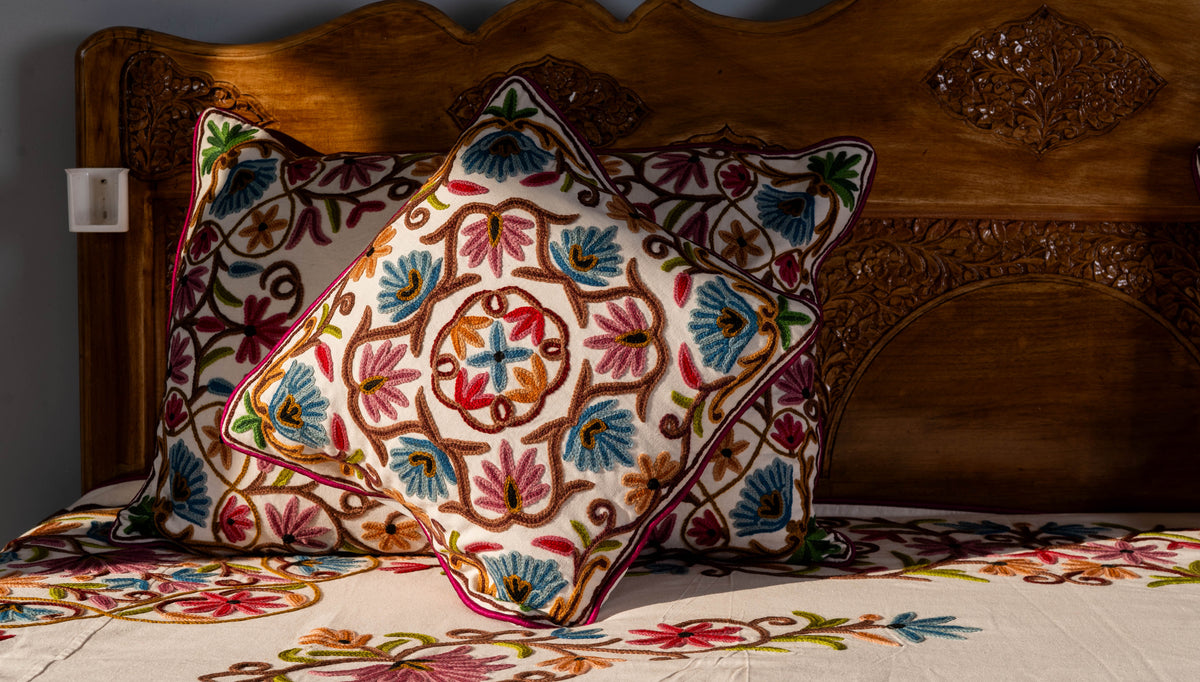 Square embroidered cushion leaning against a matching rectangular pillow on the cream floral bed cover.