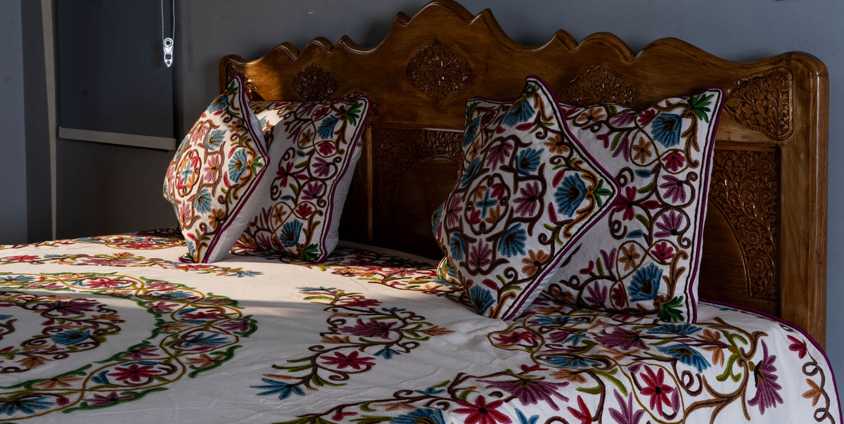 Matching crewel pillows and square cushions arranged against a carved wooden headboard above the floral bed cover.