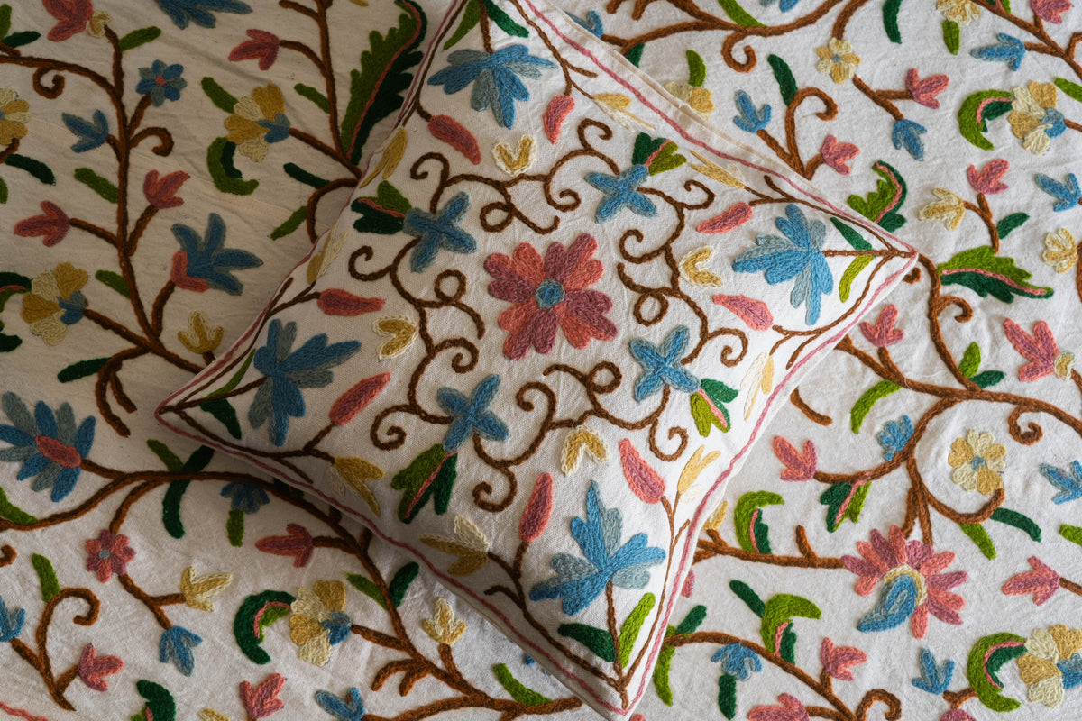 Close view of a cream cushion with pink and blue Crewel flowers on brown curling stems, resting on the matching bed cover