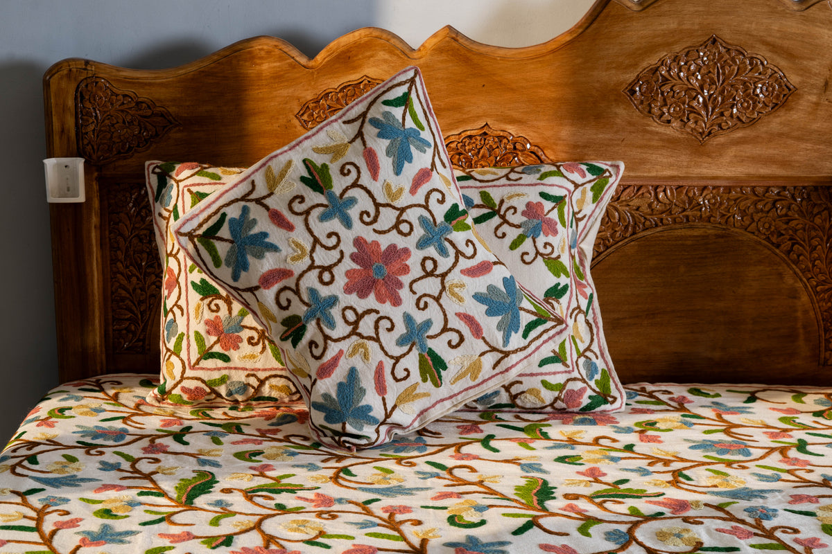 Square Crewel floral cushion in front of a matching rectangular pillow on a cream bed cover with colourful flowering vines