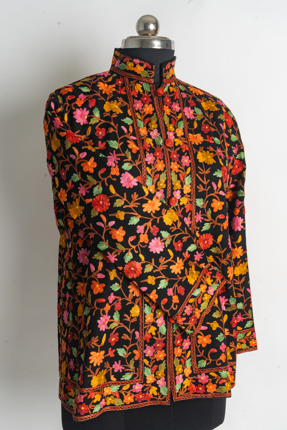 Angled front of a black short jacket with colourful Aari floral vines, red buttons and one sleeve crossed