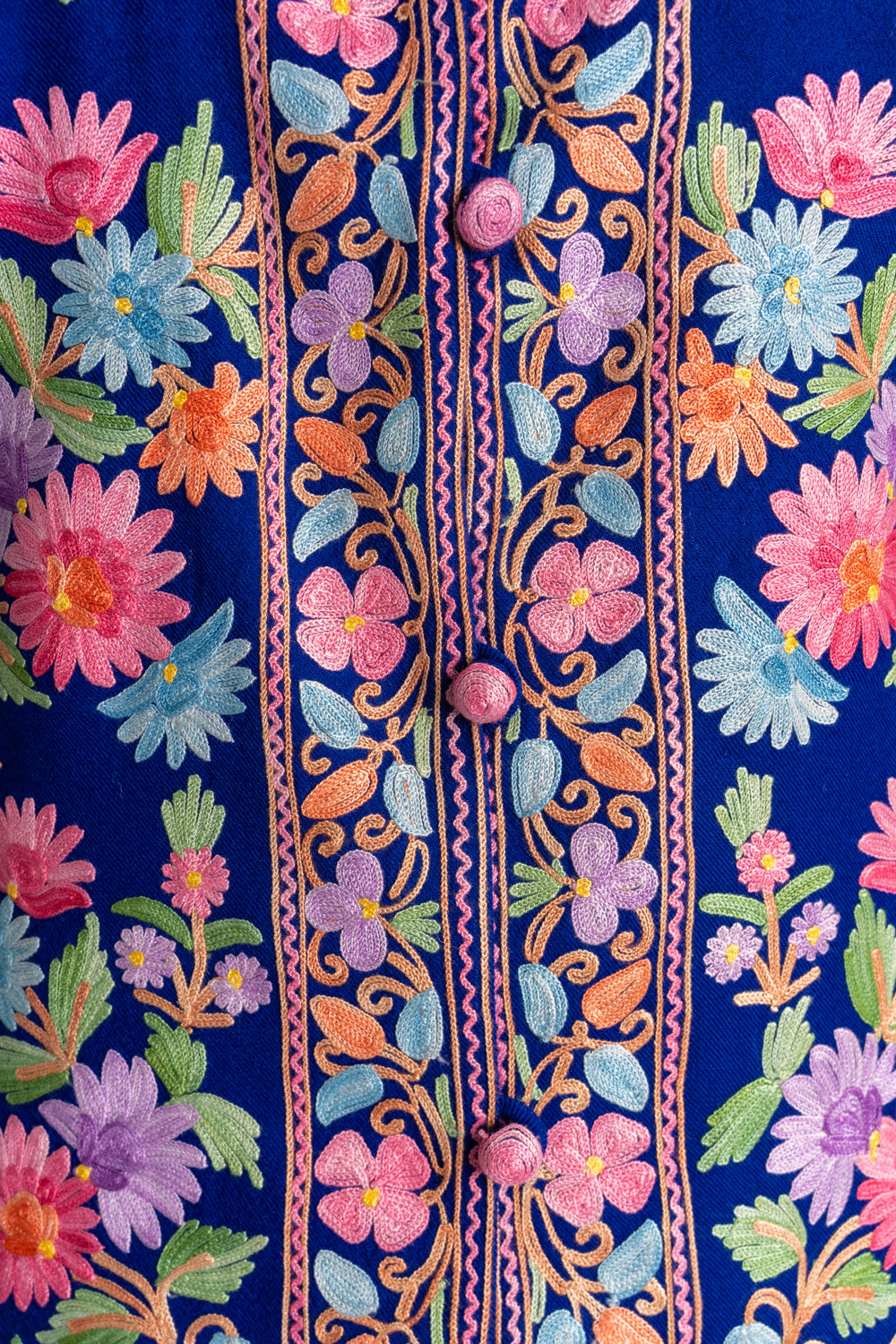 Close-up of the blue jacket’s floral front borders and pink embroidered buttons, surrounded by colourful flowers and leaves.