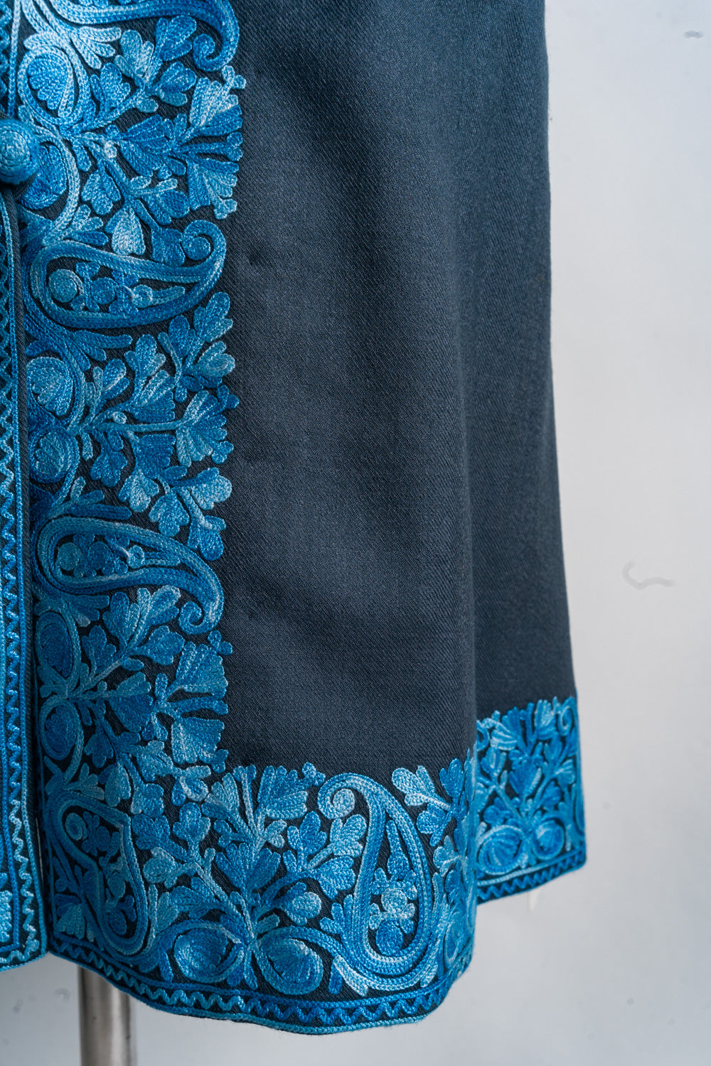 Lower corner detail of the grey jacket with blue paisley embroidery framing the front opening and hem.