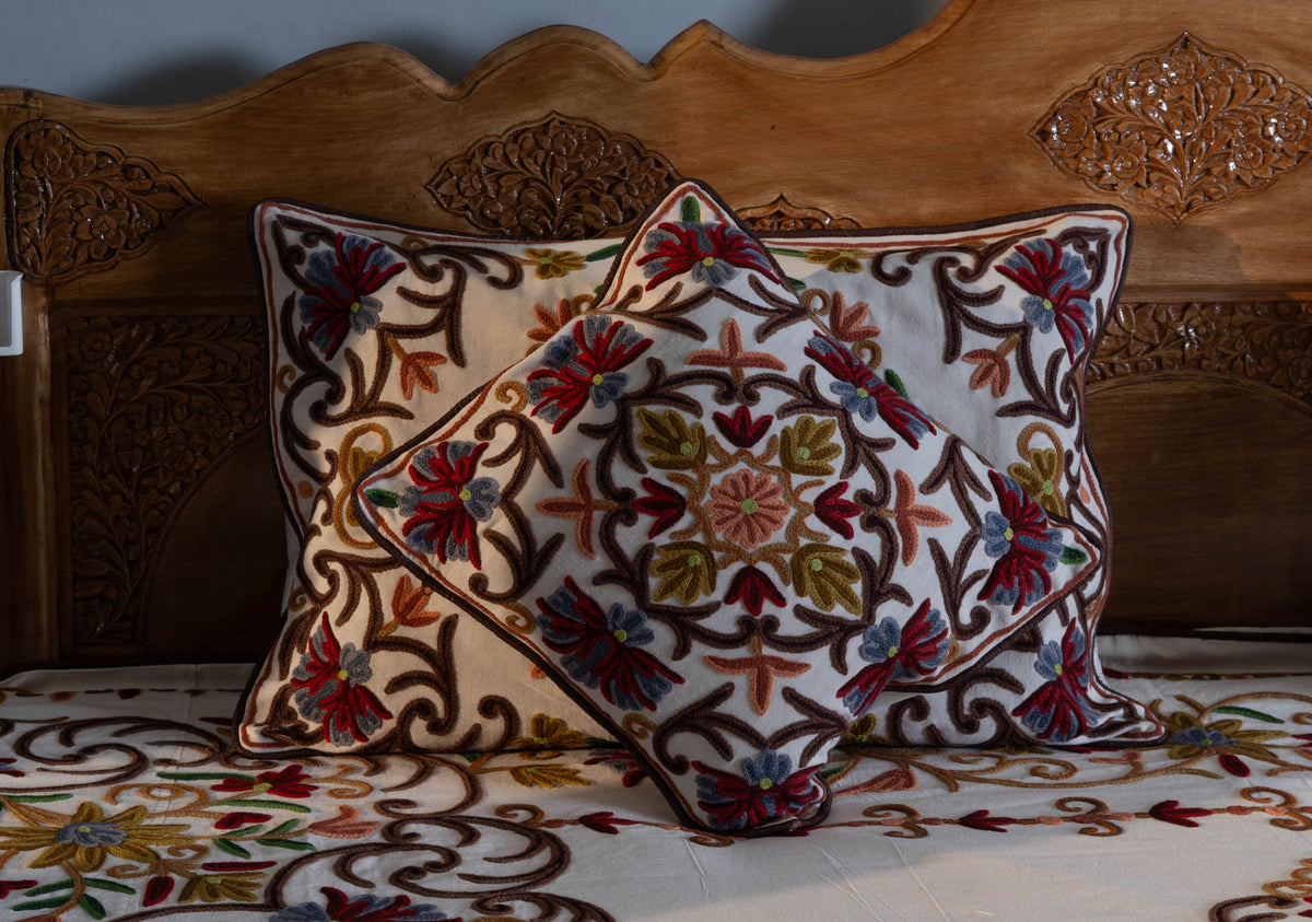 Close-up of a cream square cushion in front of a rectangular pillow, both embroidered with colourful flowers and brown vines