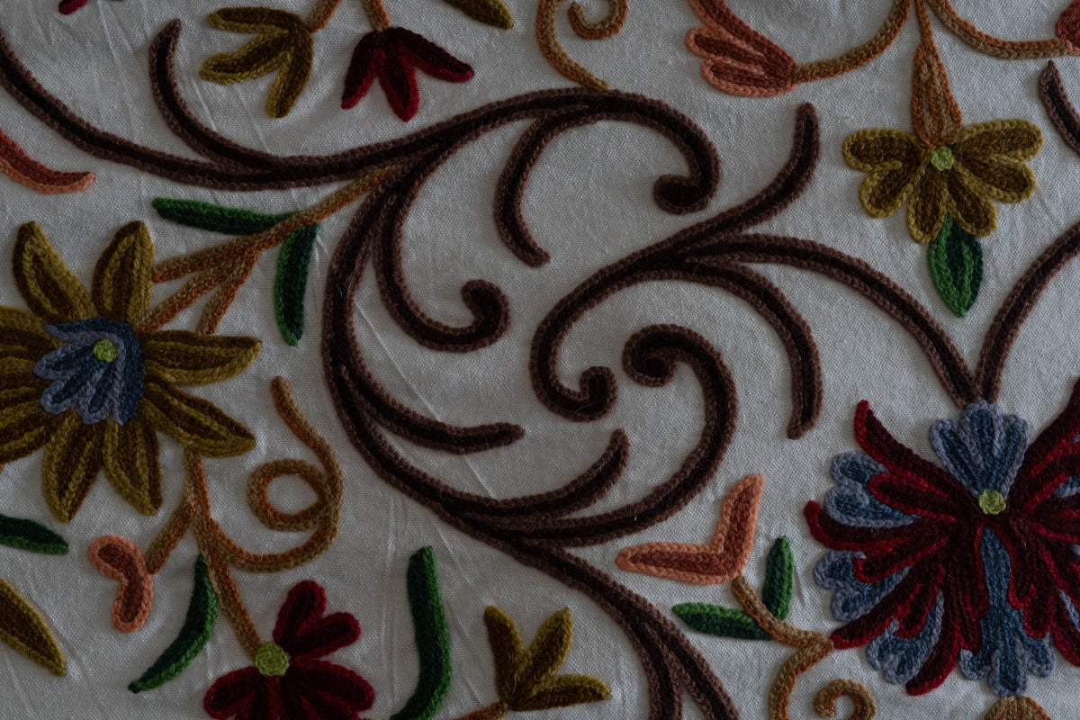 Close-up of raised Crewel stitching forming brown scrolls, red-and-blue flowers and green leaves on cream cotton