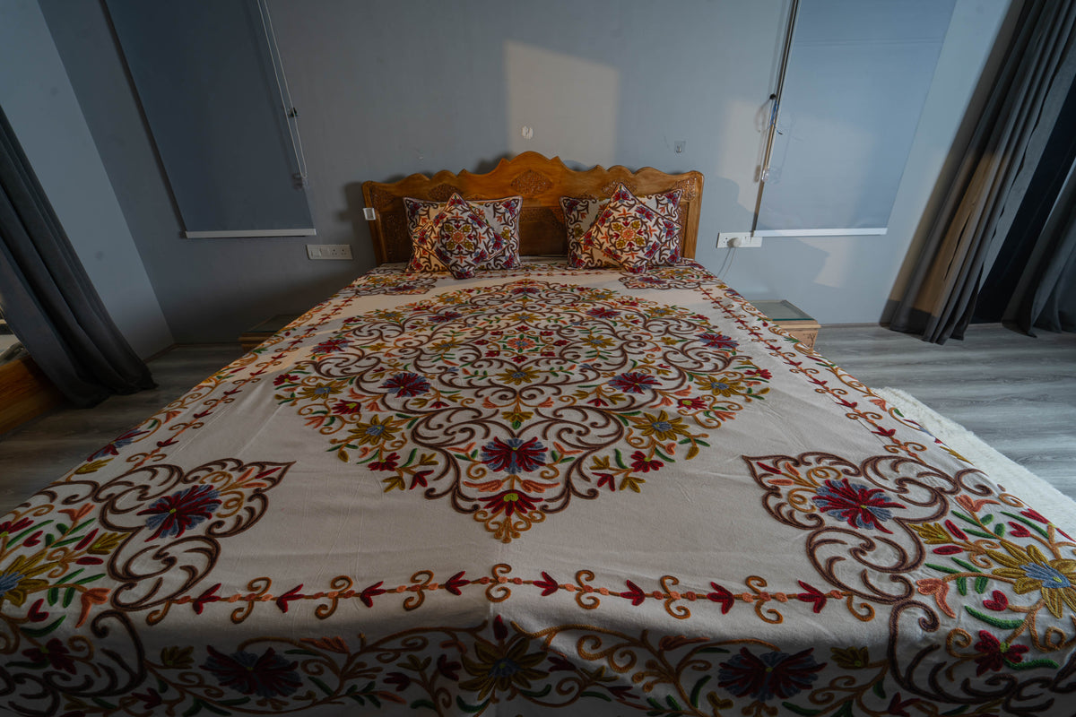 Full bed viewed from the foot, with a cream embroidered cover, central floral medallion, corner motifs and matching pillows