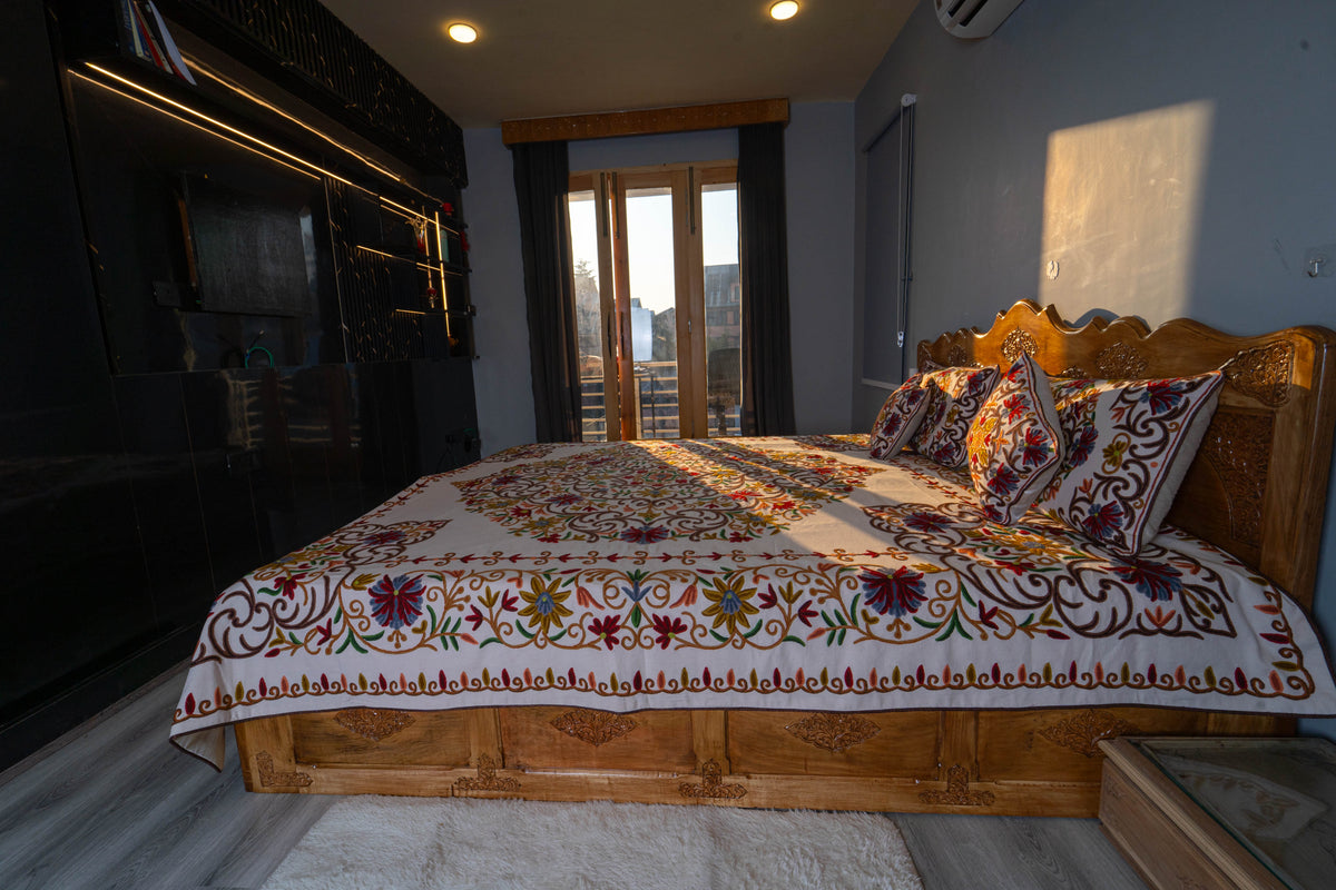 Side view of the wooden bed dressed in a cream Crewel cover with floral borders and coordinating pillows and cushions