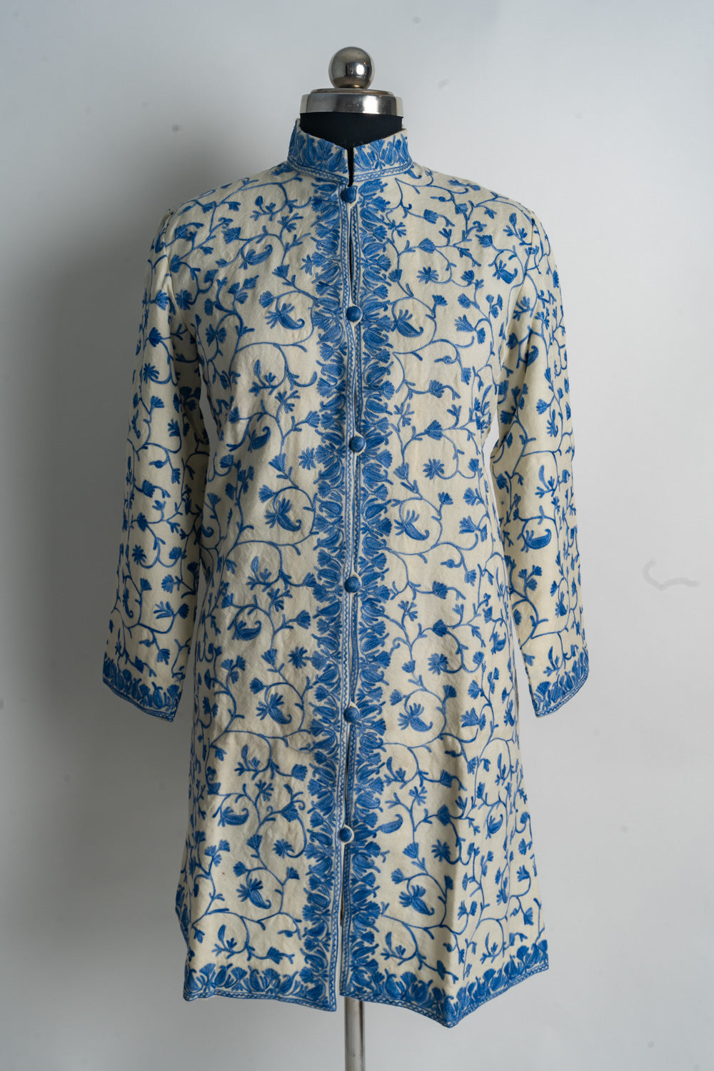 Full front view of a cream jacket covered in delicate blue floral vines, with a stand collar and narrow embroidered borders.