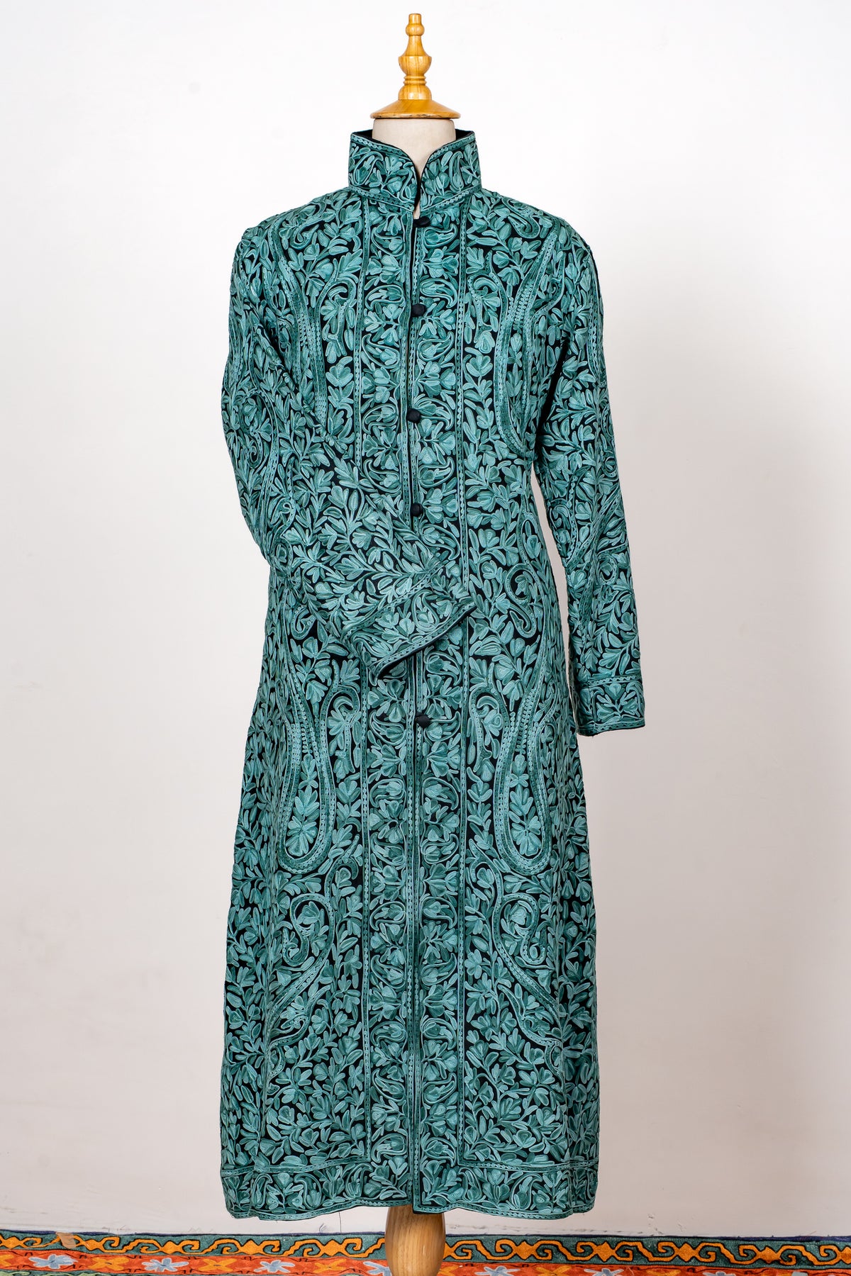 Front view of a teal long jacket covered in turquoise Aari floral vines, with a high collar and one sleeve crossed