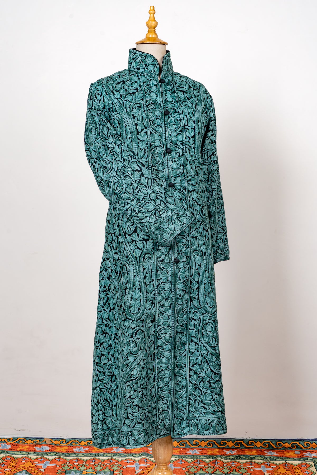 Full-length teal jacket on a mannequin with all-over turquoise Aari embroidery and one sleeve folded across the front