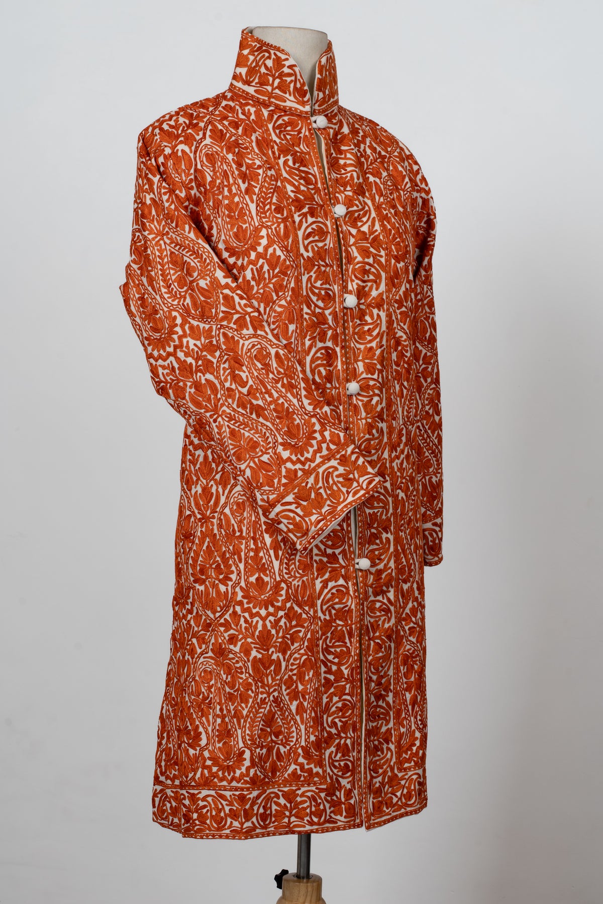 Angled full view of the orange-and-white embroidered jacket, showing its fitted waist, crossed sleeve and long silhouette.