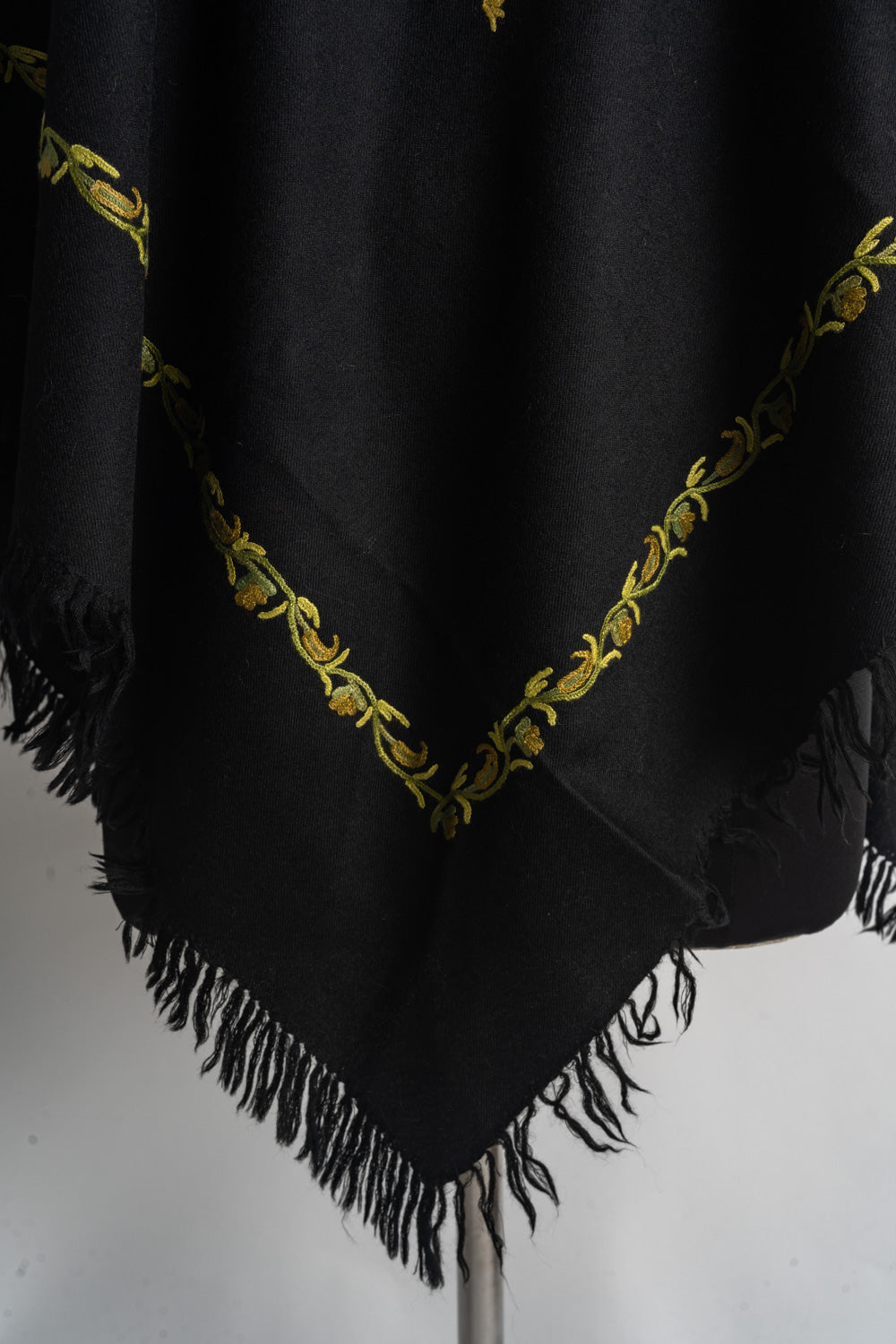 Pointed corner of a black poncho with a golden yellow and green Aari vine border above black fringe