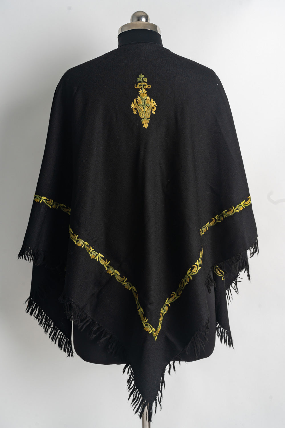 Back of a black poncho with a small golden embroidered motif, a narrow vine border and a pointed fringed hem