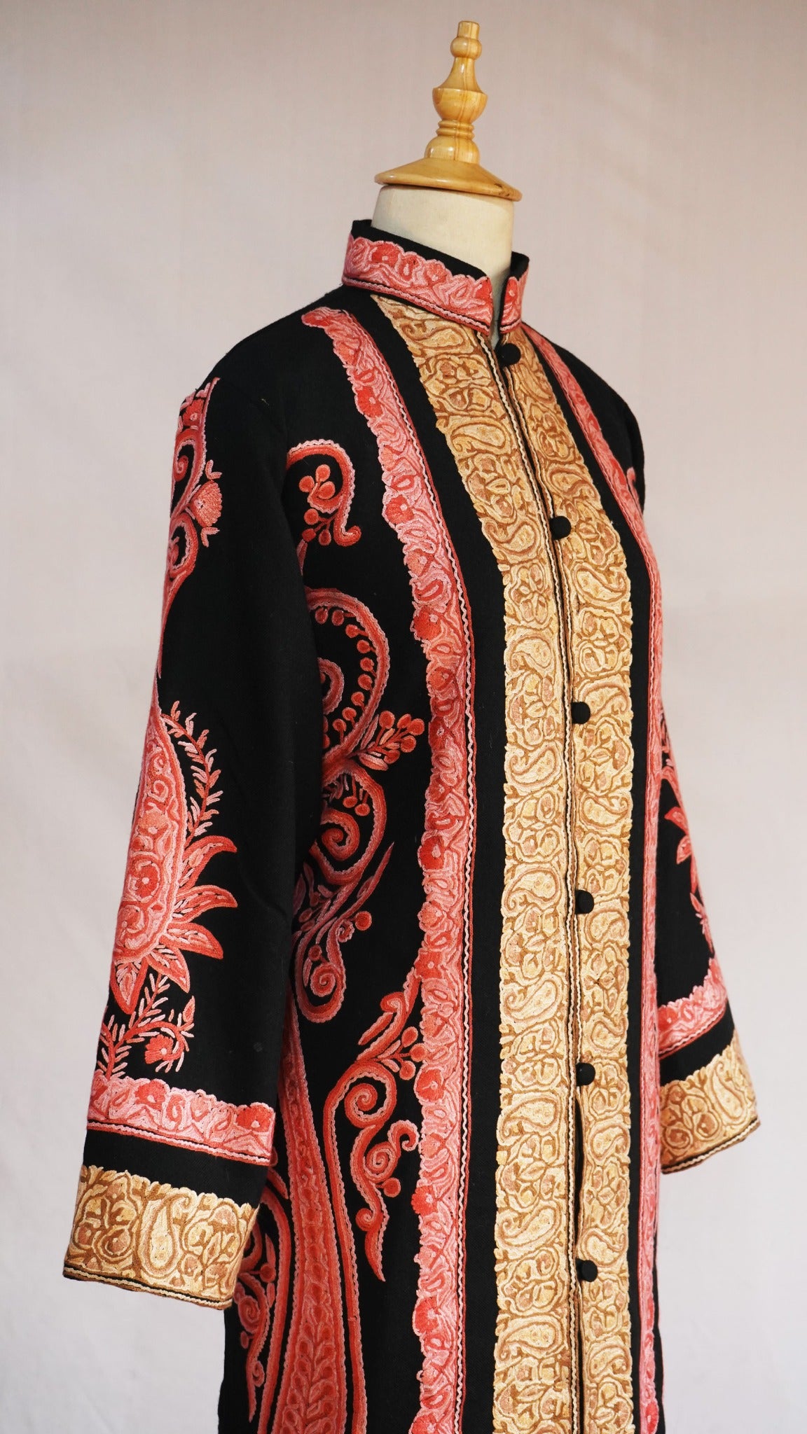 Angled front of the black jacket with a high collar, golden embroidered opening and coral-pink paisley motifs on the sleeve and side
