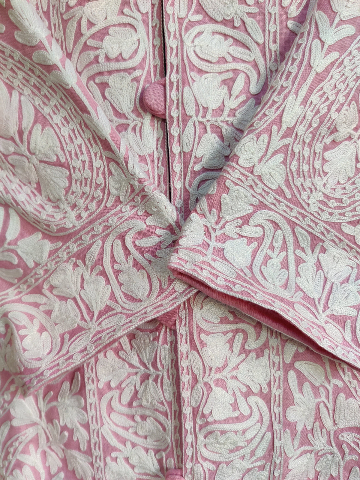 Close-up of the pink jacket’s crossed cuffs and covered buttons with dense white floral and paisley embroidery.