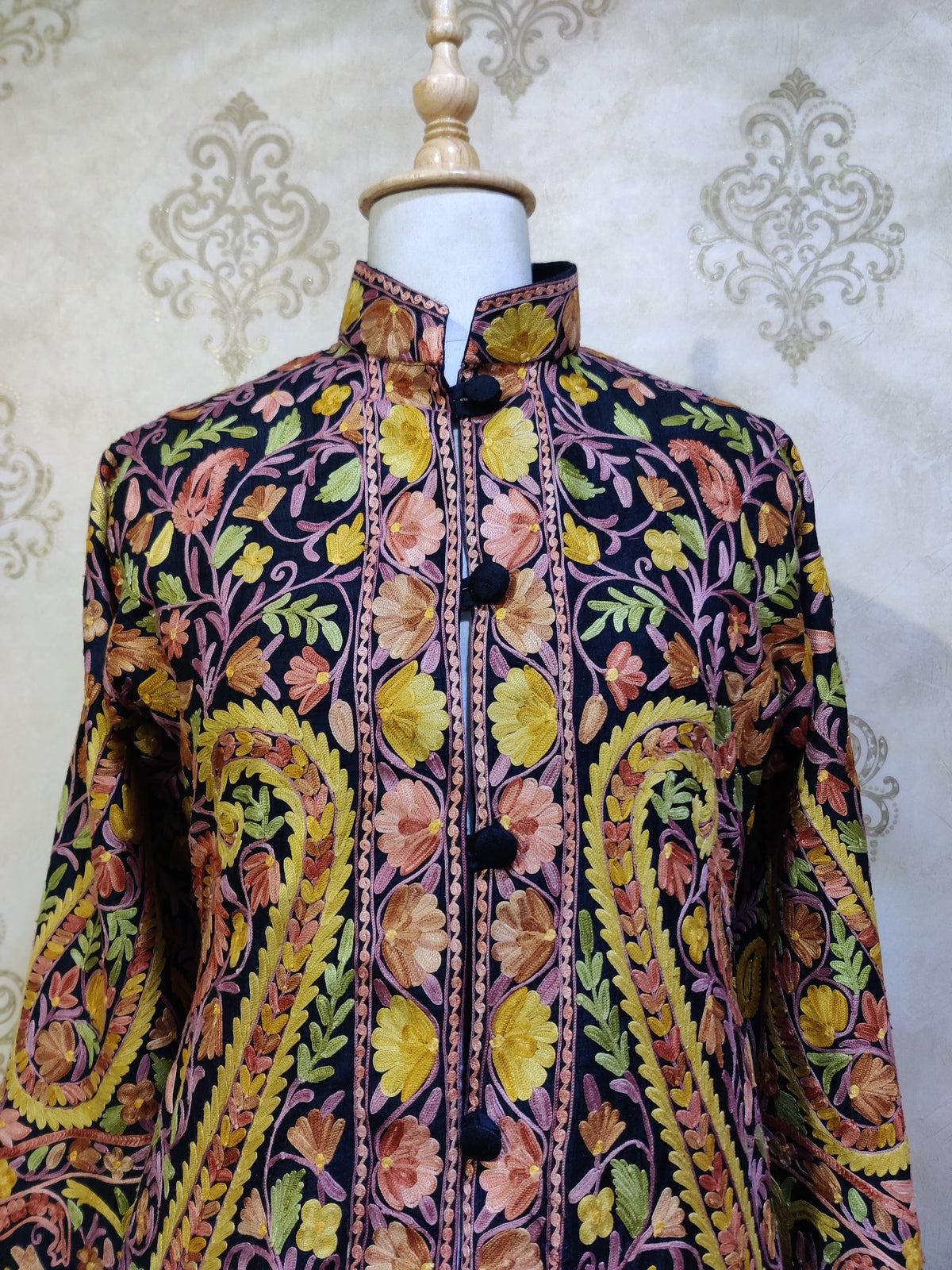 Upper front detail of a dark jacket showing pink and yellow Aari flowers around the stand collar and buttoned placket