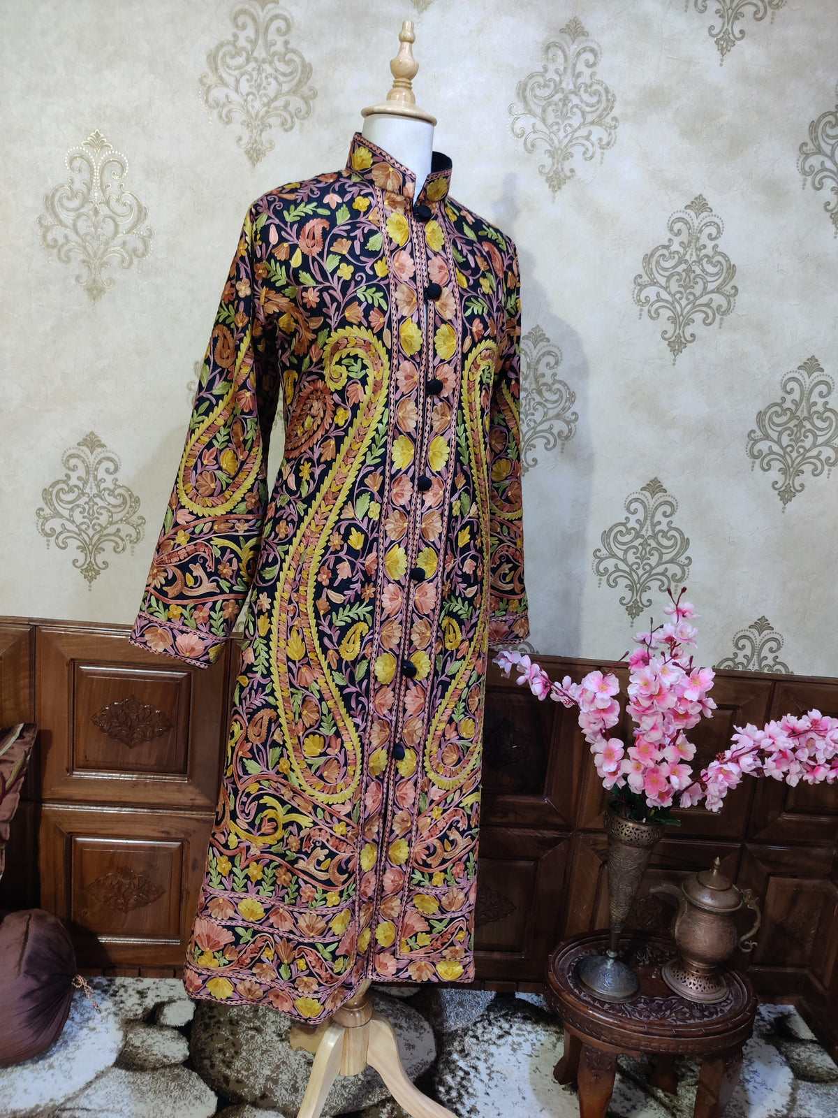 Angled full view of a dark long jacket with large yellow-green Aari paisleys and pink floral embroidery from collar to hem