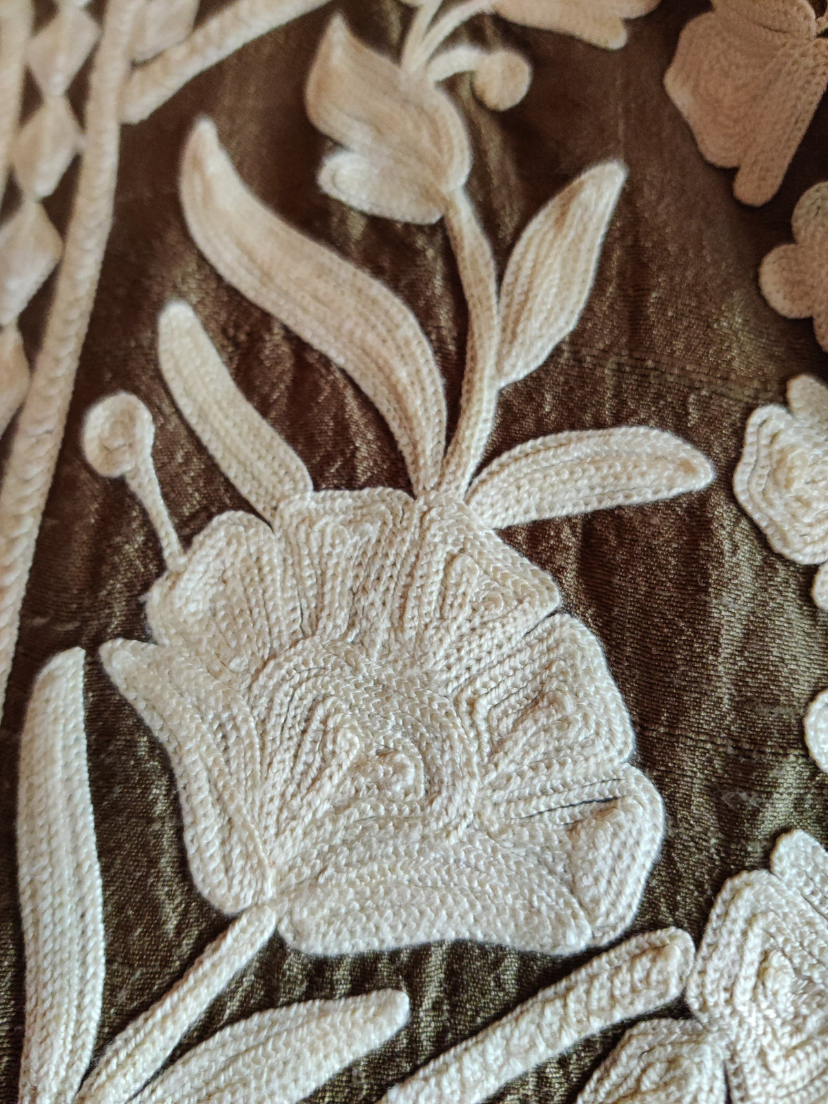 Macro detail of a white Aari flower and curling leafy stem on the brown jacket fabric.