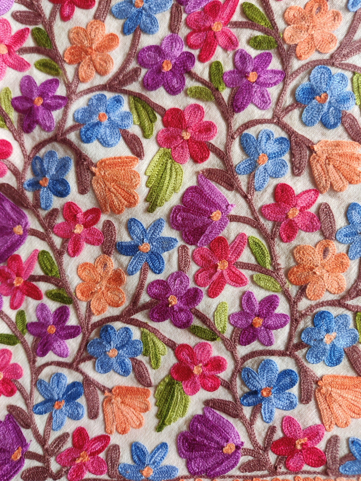 Macro detail of blue, purple and pink Aari flowers, peach buds and green leaves on white jacket fabric