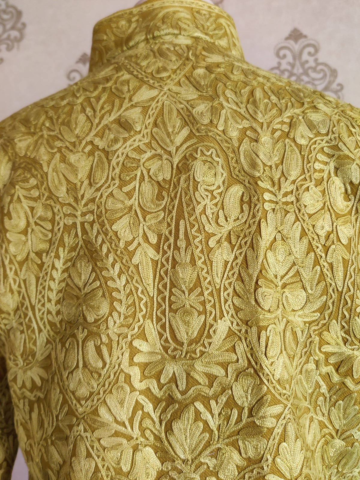 Close-up of the mustard-yellow jacket’s upper back and standing collar, showing tonal Aari paisleys, flowers and leaves.