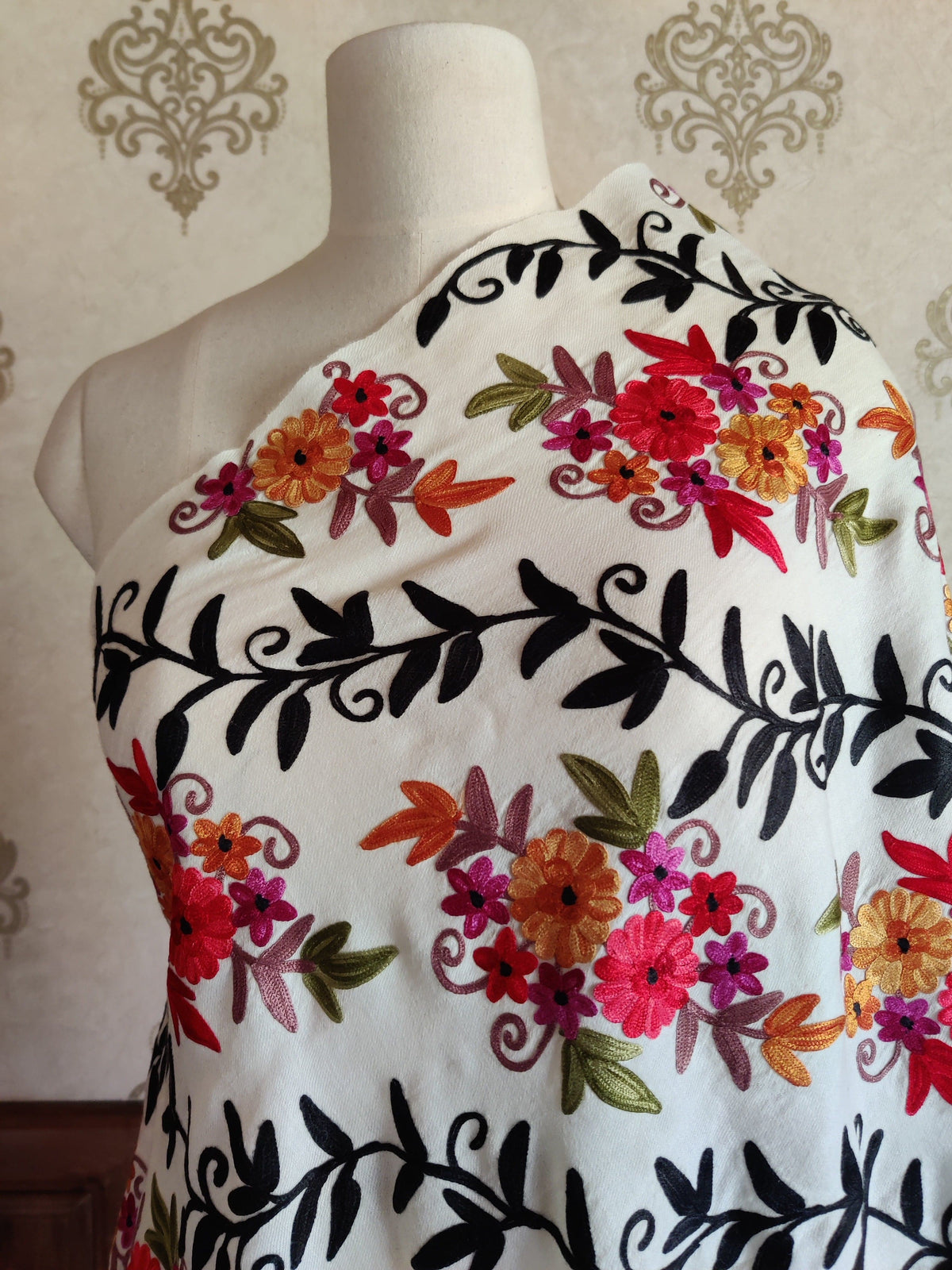 Close-up of a white merino wool stole across a mannequin's chest, showing bright Aari flowers between bold black leafy branches.