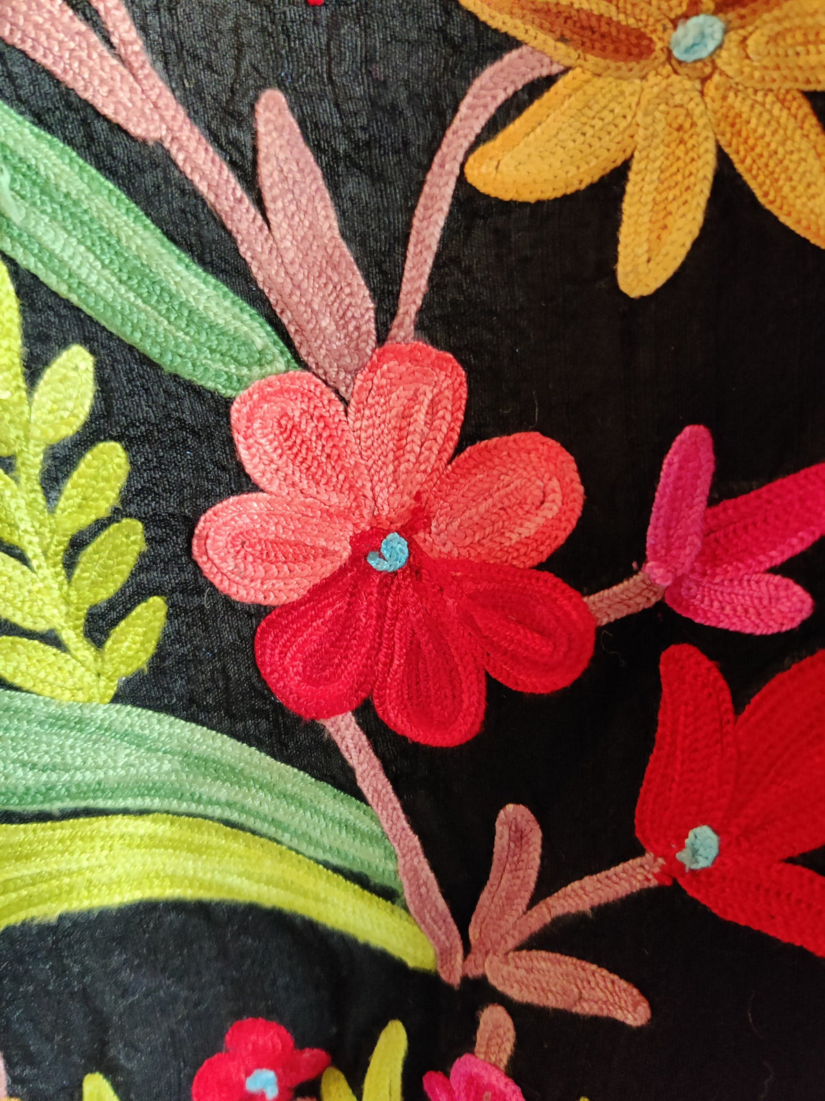 Macro view of a red and coral Aari flower with a turquoise centre and green leaves on black jacket fabric