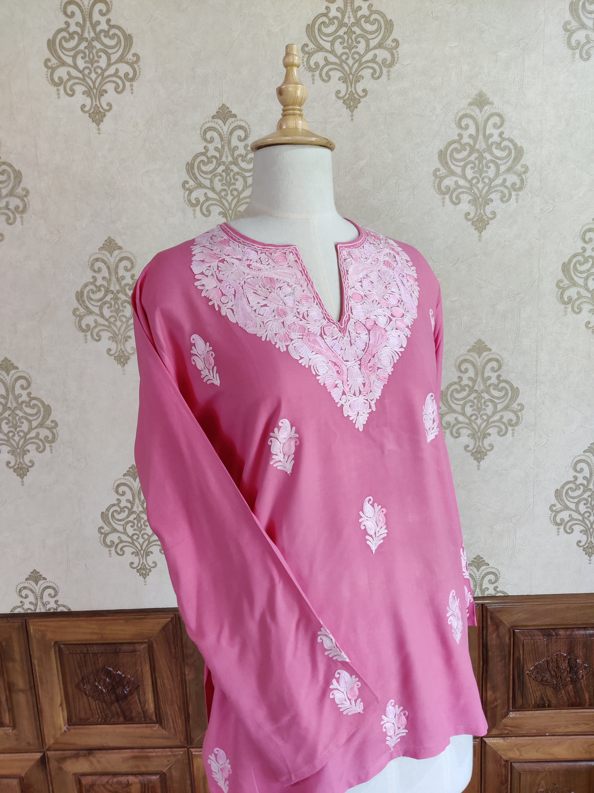 Angled view of a pink kurti with long sleeves, a white embroidered neckline and small floral motifs across the body.