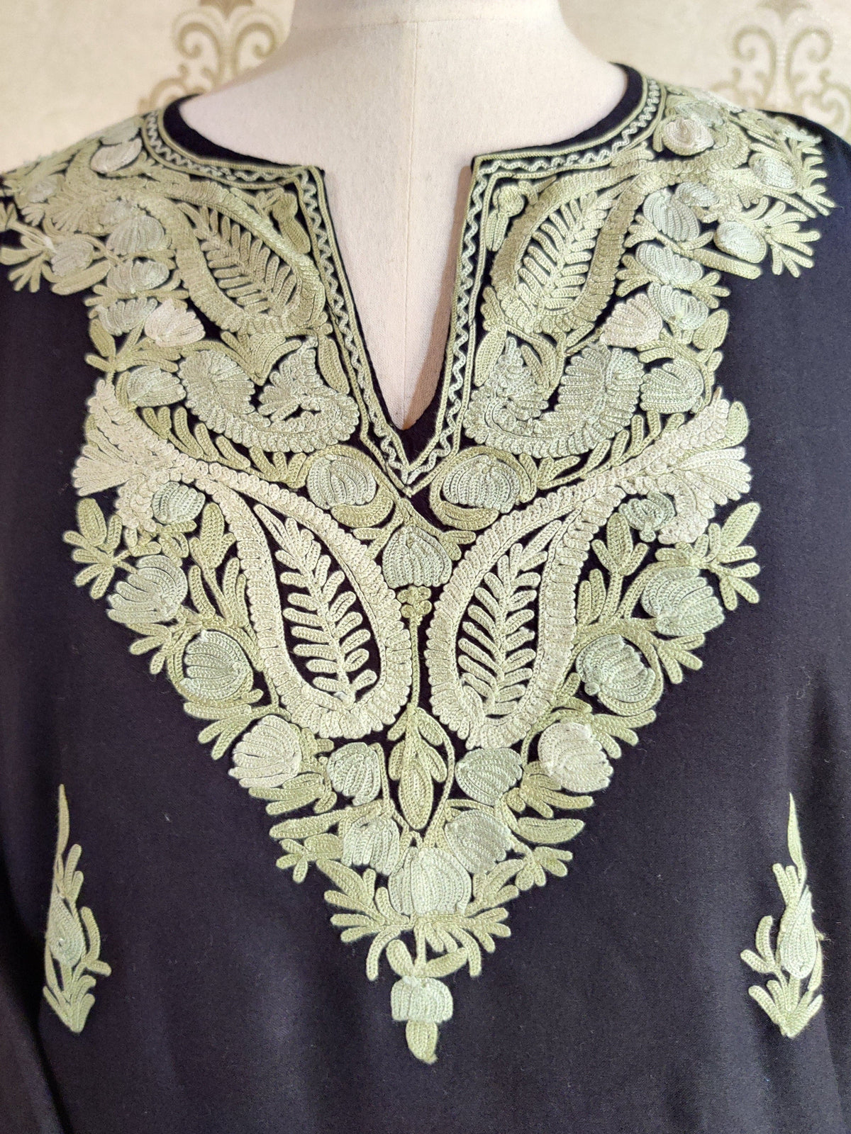 Close-up of the black kurti’s split neckline framed by mint-green Aari flowers, leaves and paired paisley shapes.