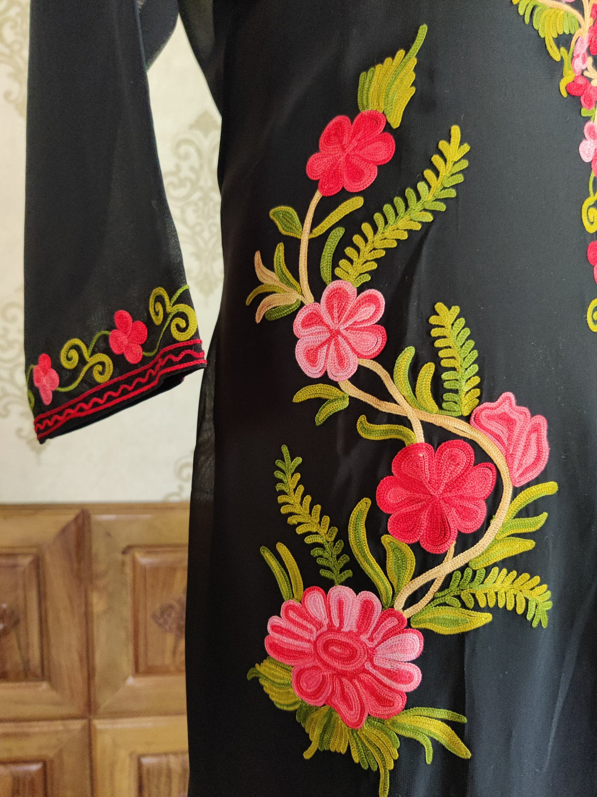 Black kaftan detail with pink and red Aari flowers on green stems, beside a floral embroidered sleeve cuff