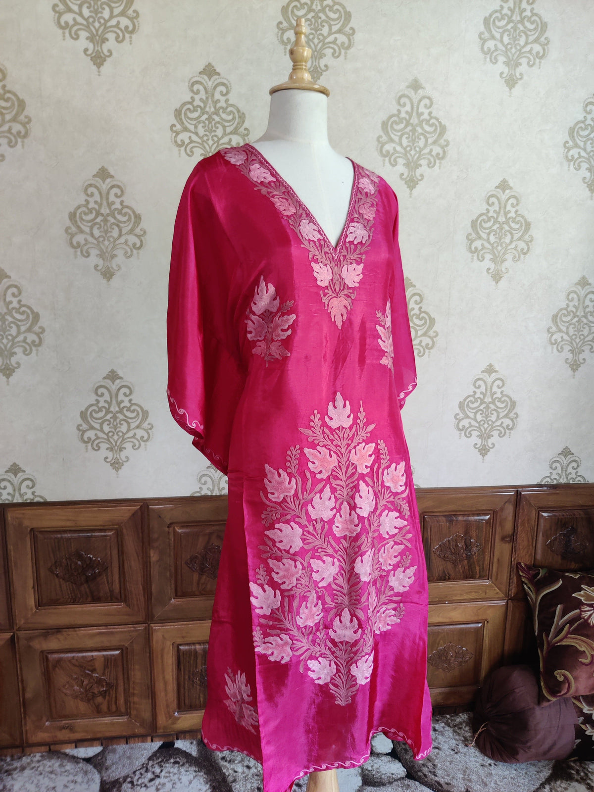 Full angled view of a long fuchsia kaftan with loose sleeves, a pale pink embroidered V-neck and a large leaf motif down the lower front
