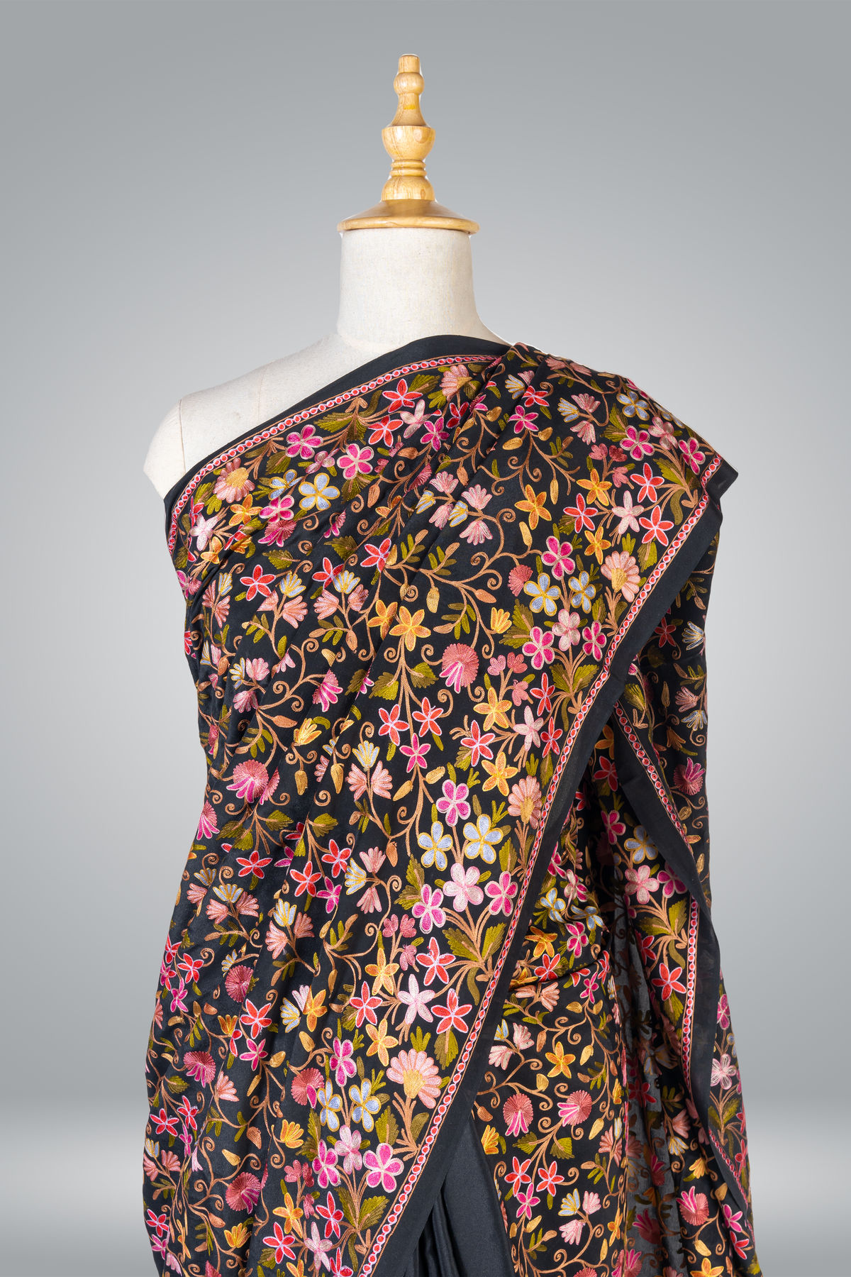 Closer draped view of the black crepe saree on a mannequin, with multicoloured Aari flowers and a narrow pink-edged border.