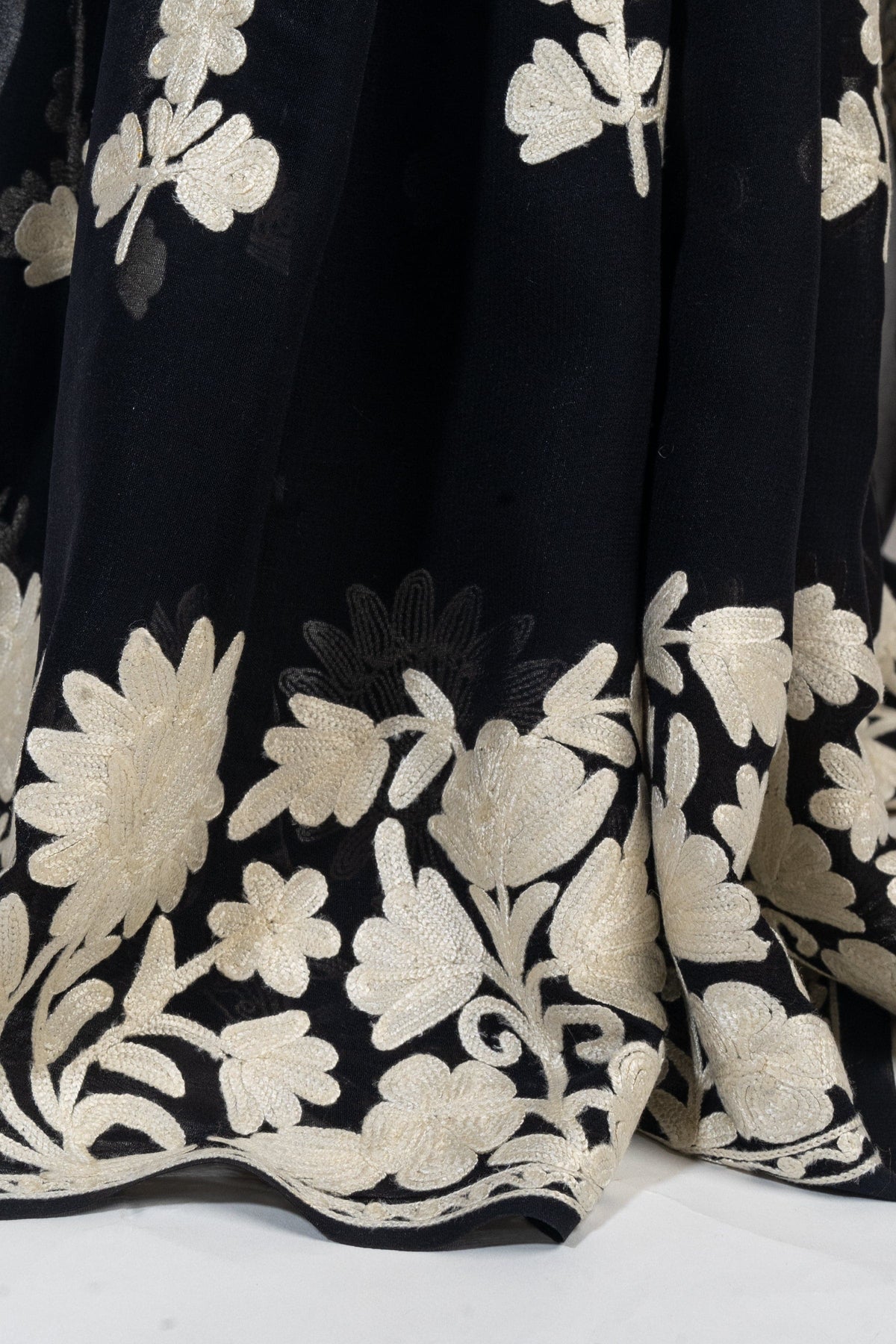 Close-up of cream Aari flowers and leaves along the black crepe saree’s lower border, shown in soft folds.