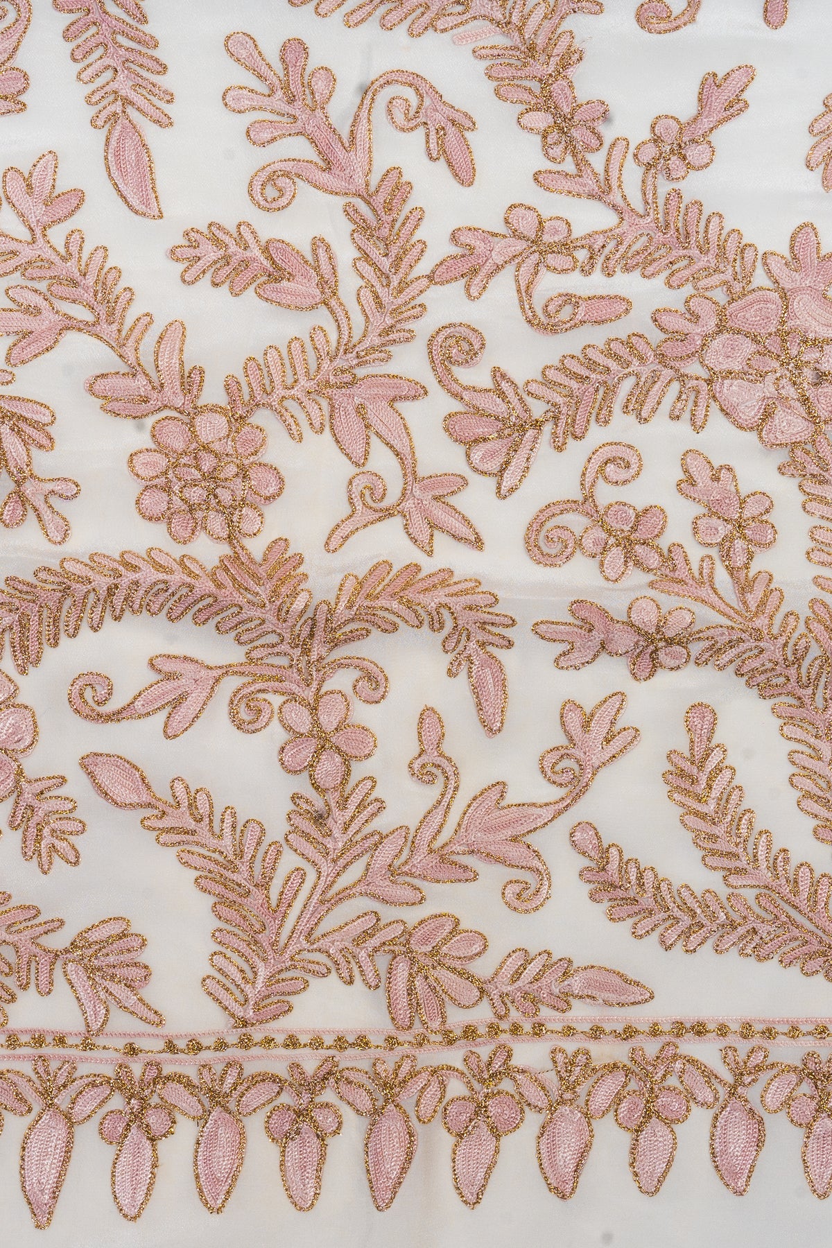 Close-up of pale beige-pink Aari flowers and curling leaves outlined in gold Zari, above a hanging-leaf border on white