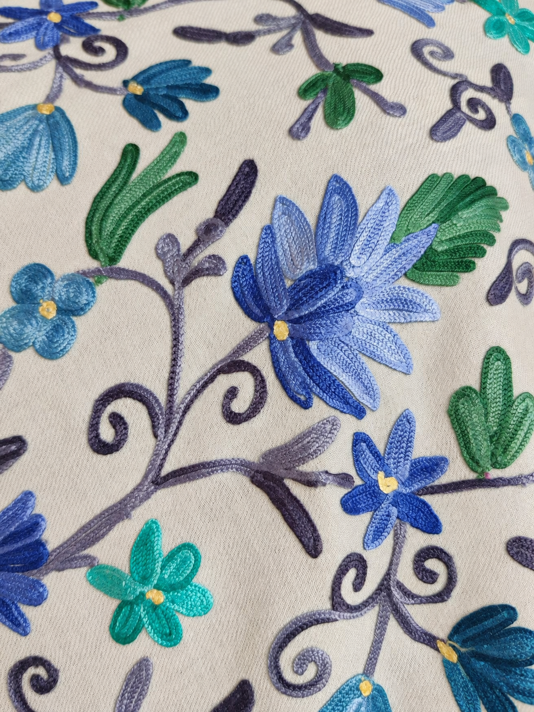 Close-up of blue and turquoise Aari floral embroidery and purple trailing vines on ivory fine Merino wool