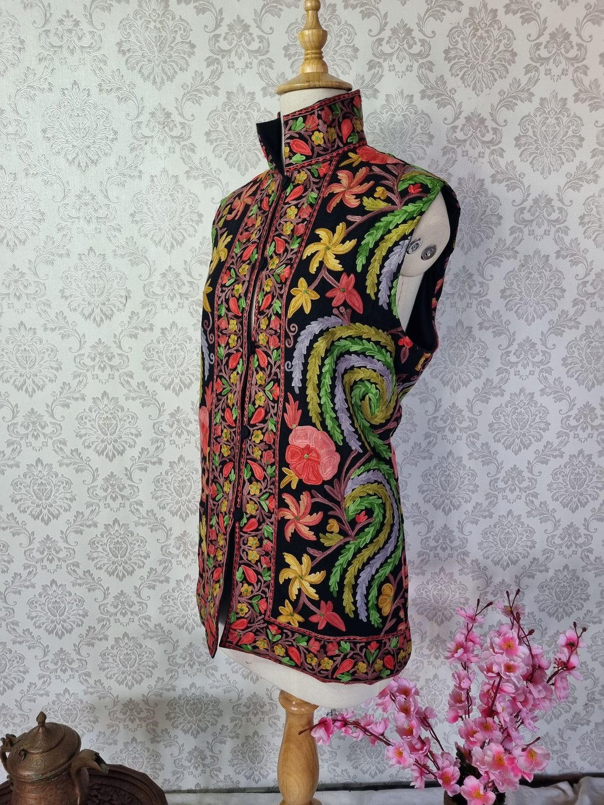 Three-quarter view of a black sleeveless jacket on a mannequin, with bright Aari floral motifs and curling green-and-lavender paisleys.