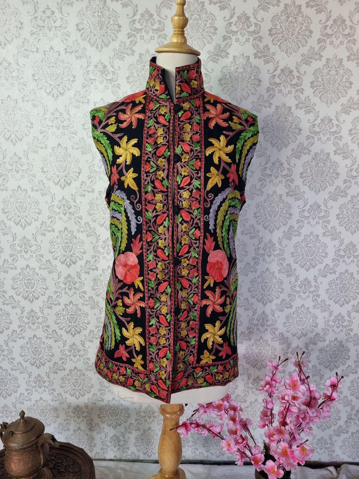 Front of a black sleeveless jacket on a mannequin, with green paisleys, red and yellow Aari flowers, a stand collar and floral hem border.