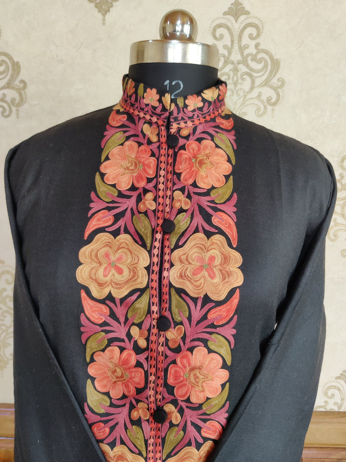 Upper front of a black jacket on a mannequin, with large peach and coral Aari flowers along the placket and a floral stand collar.
