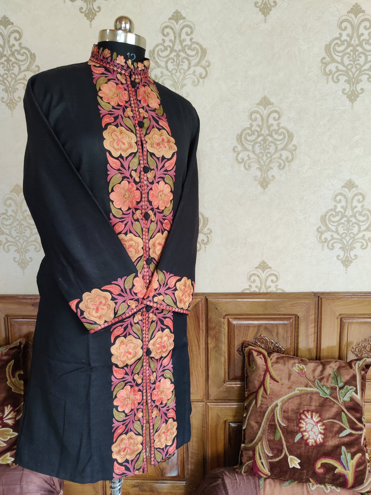 Angled front of a black long jacket with crossed sleeves, showing a peach-and-coral Aari floral panel and matching cuffs.