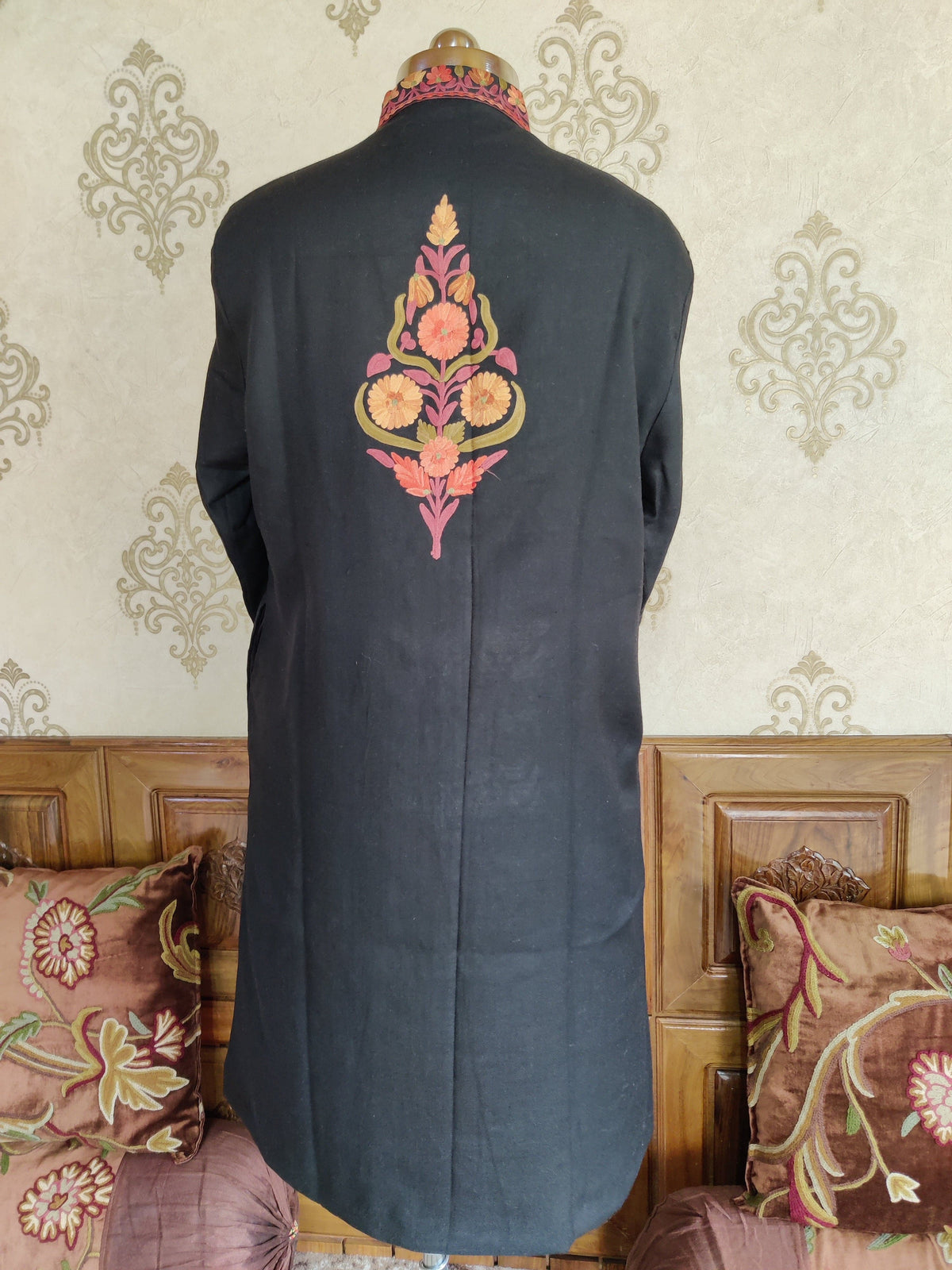 Back of a black long jacket on a mannequin, with a single peach, coral and olive-green Aari floral motif between the shoulders.