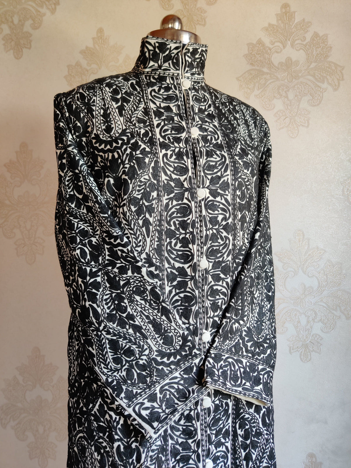 Angled upper view of the black-and-ivory jacket, highlighting dense paisley embroidery, crossed cuffs and the buttoned front.