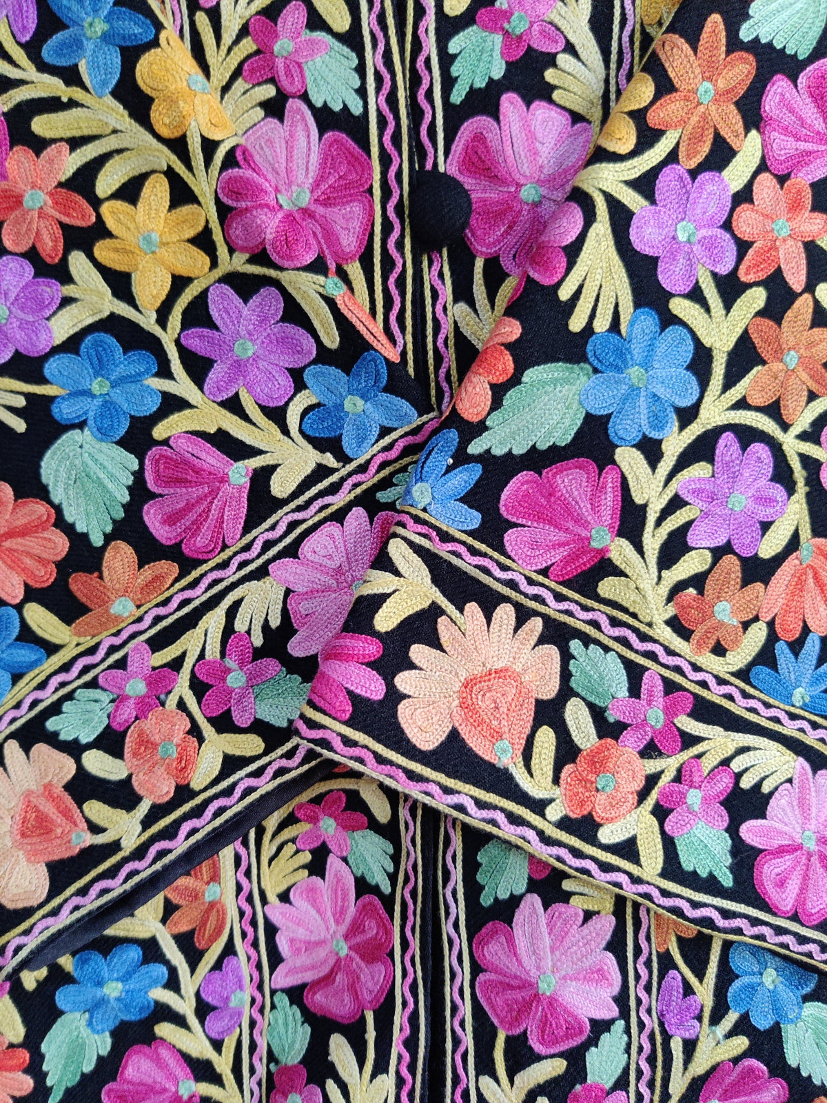Close-up of pink, purple, blue and peach Aari flowers around a black button and crossed floral cuffs.