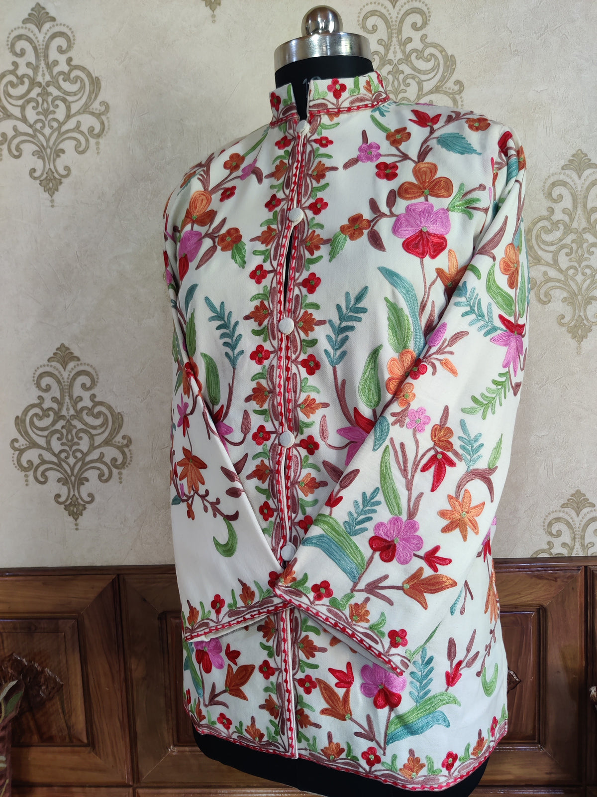Angled view of a short ivory floral jacket with crossed sleeves, a stand collar and narrow red-edged borders.