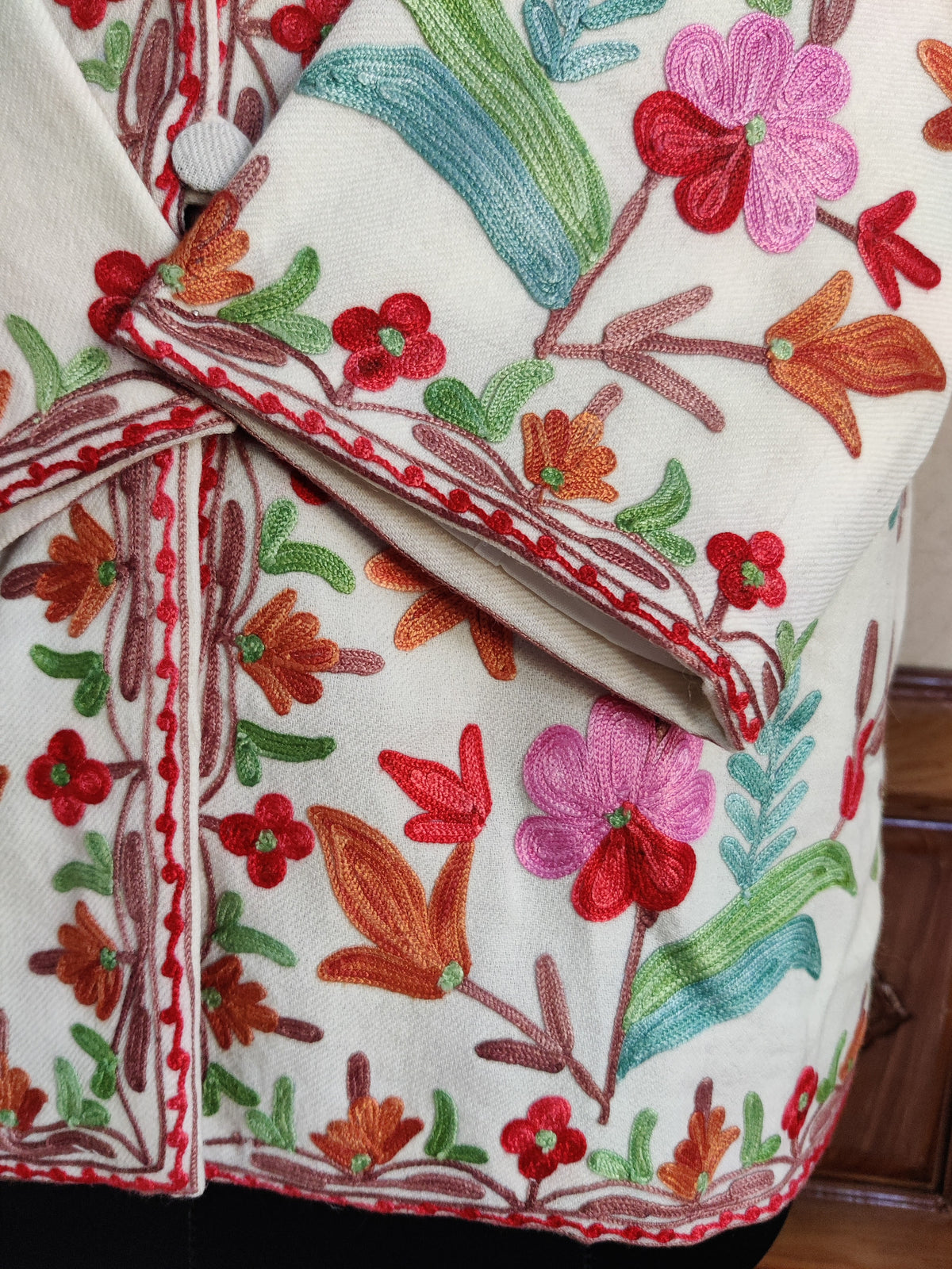 Close-up of an embroidered ivory cuff across the front, showing pink and red flowers, orange buds and teal-green leaves.