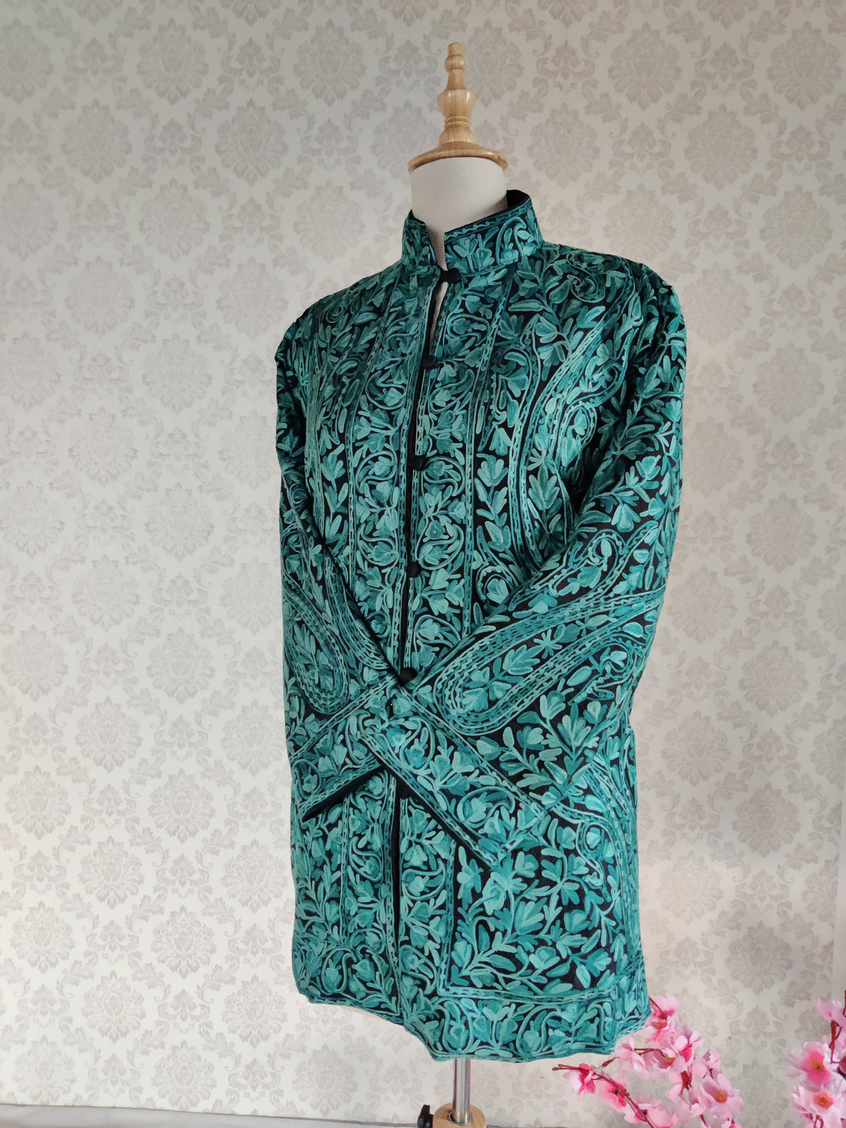 Angled front of a black jacket with crossed sleeves, showing teal Aari paisleys, dense floral vines and a matching buttoned placket.
