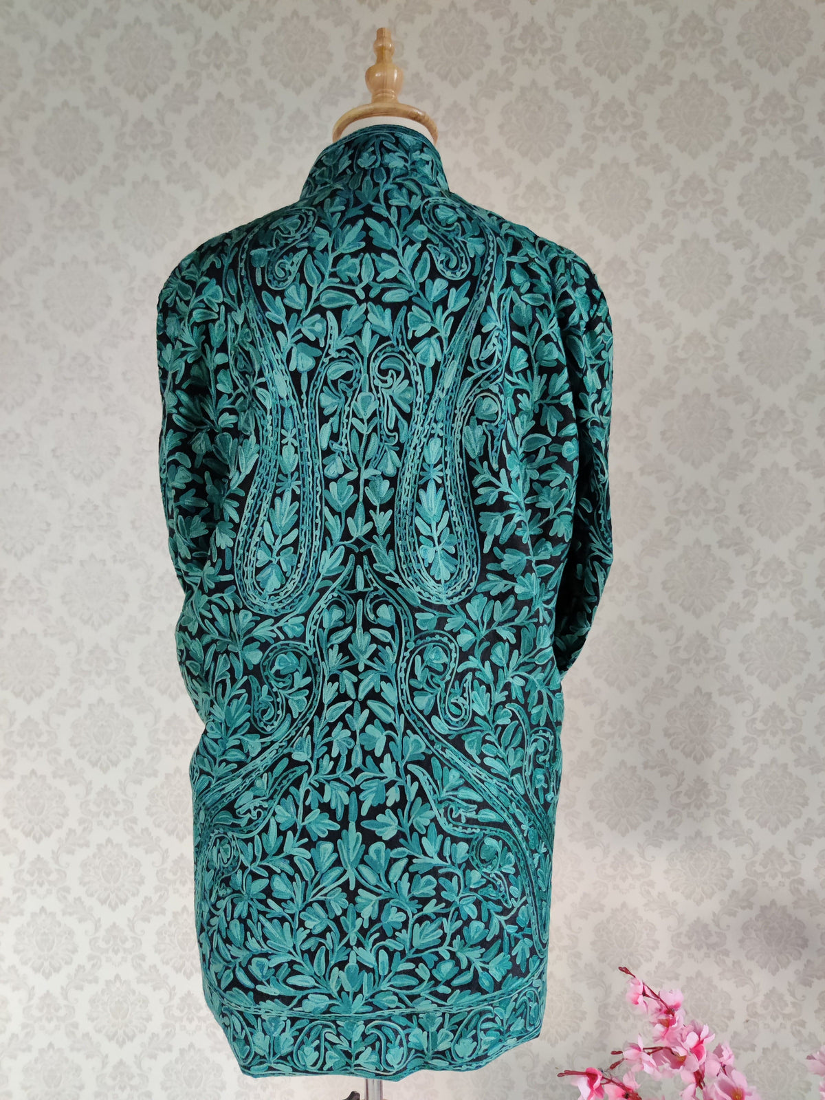 Back of a black jacket on a mannequin, with large teal Aari paisleys among dense leafy vines and a matching floral hem border.
