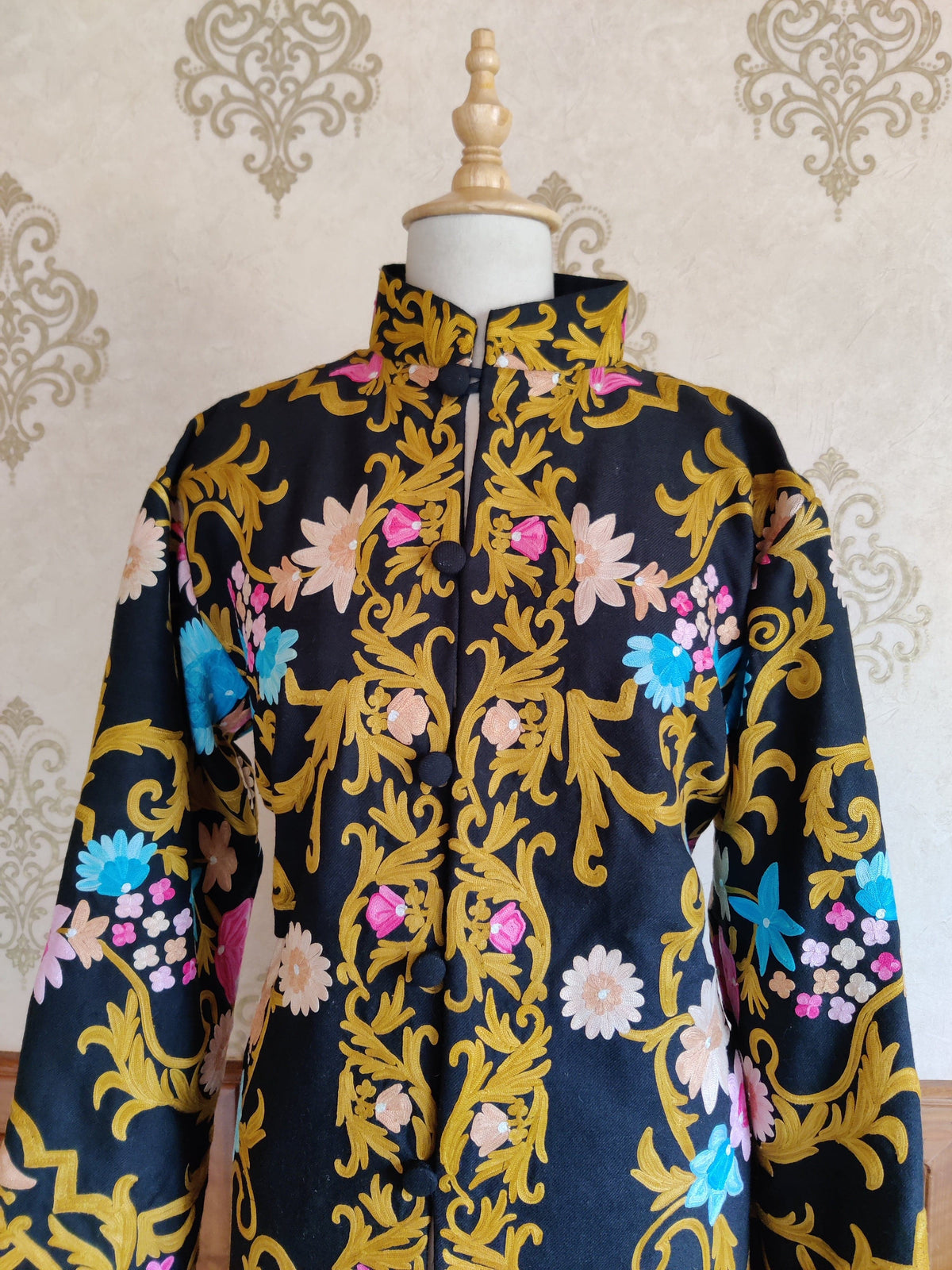 Upper front of a black coat with golden Aari leafy vines, pink flower buds and blue and beige blossoms beside the buttoned opening